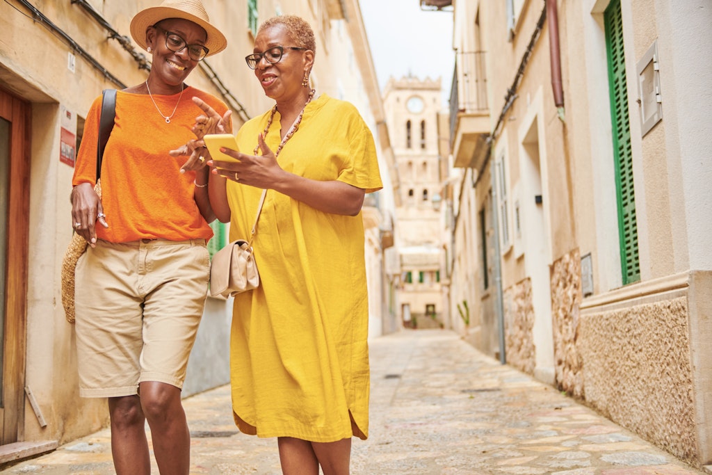 Using a mobile app on their smart phone to guide their way around a traditional old town in Majorca Spain.
1438234950
Two older black women walking along a cobbled street in Spain while looking at their smartphone and laughing