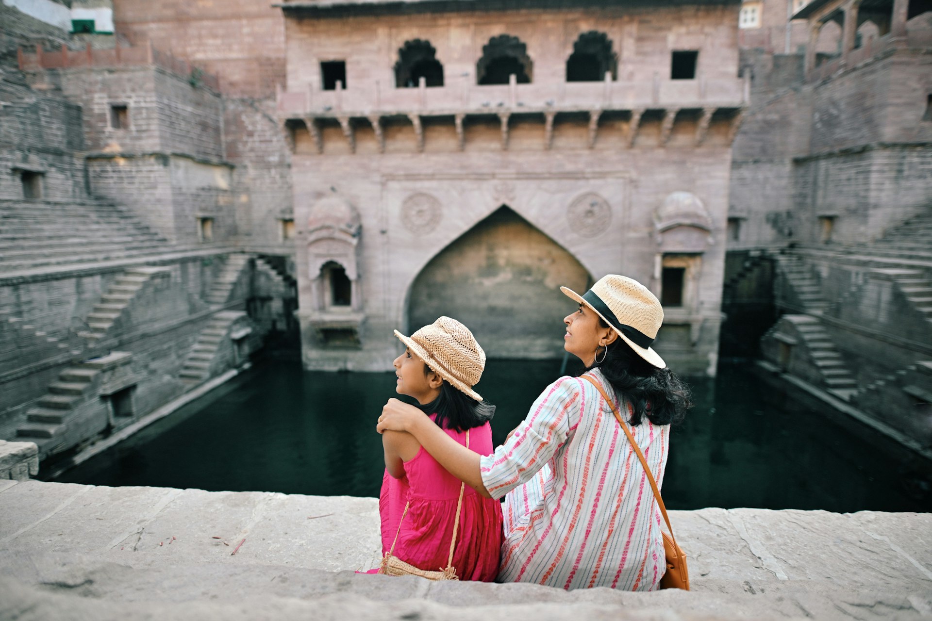 Rear view of tourist mother and daughter wearing a hat sitting at a step well (Toorji Ka Jhalra) in Jodhpur, Rajasthan