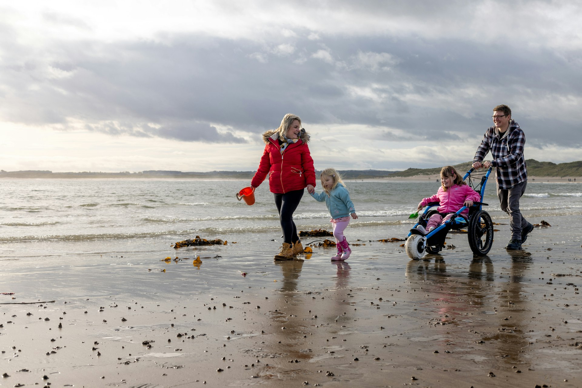 A family of four on an English beach on a slightly grey day. The mum and youngest walk along with a bucket and spade; the dad pushes the elder child in a beach-adapted wheelchair