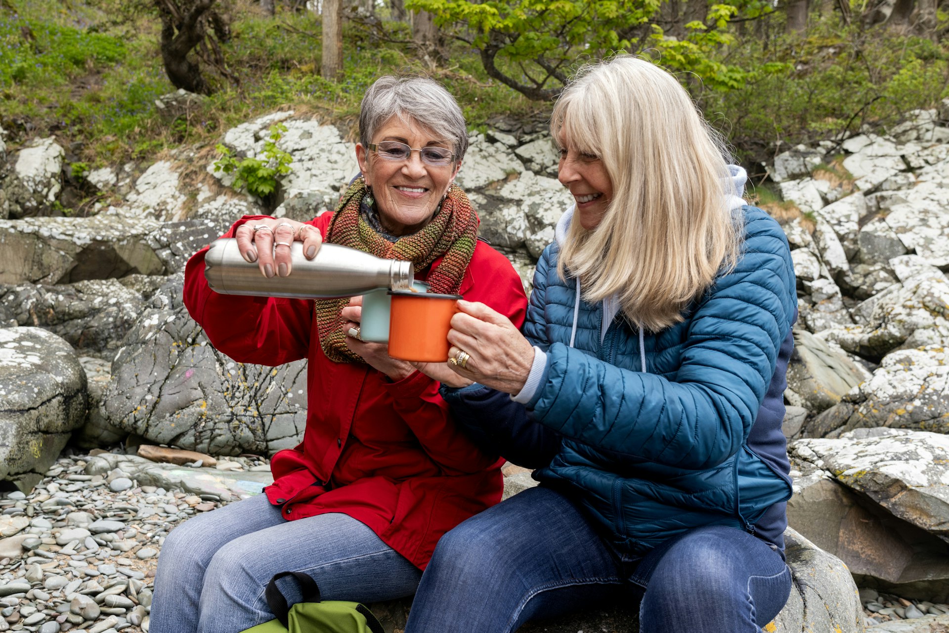 Two women taking a break on a hike to sit on a rock and drink from a thermos of water