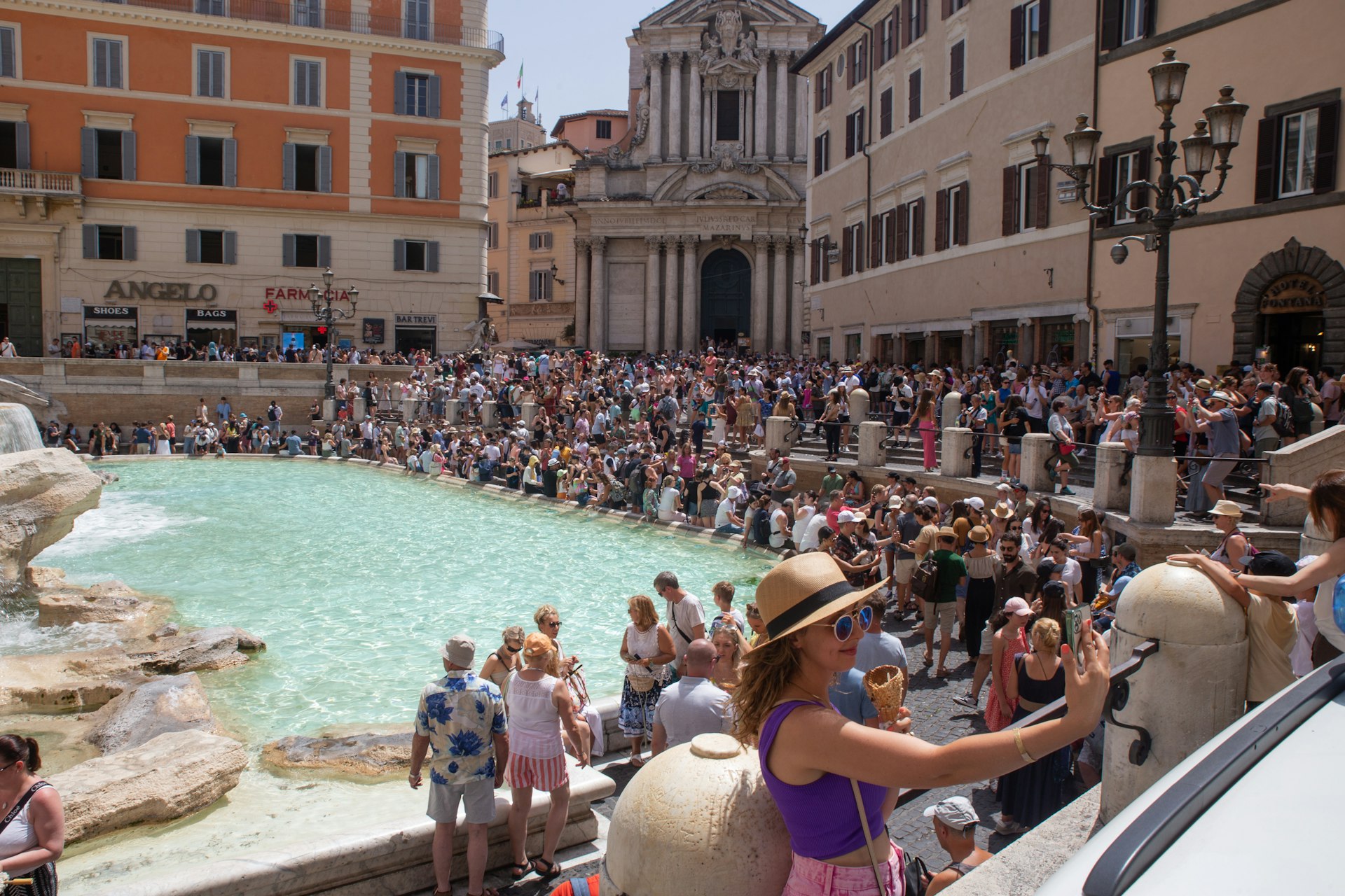 A woman takes a selfie with an ice cream in front of Trevi Fountain on a hot summer day