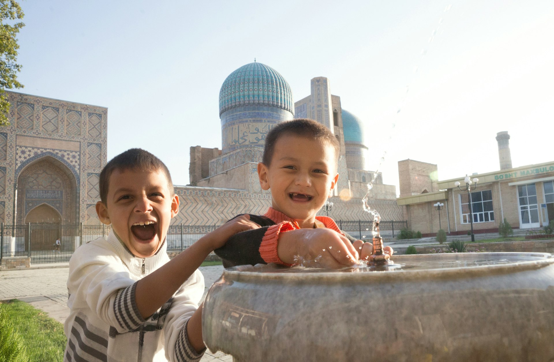 Two boys splash in a small water fountain in front of a domed and tiled mosque