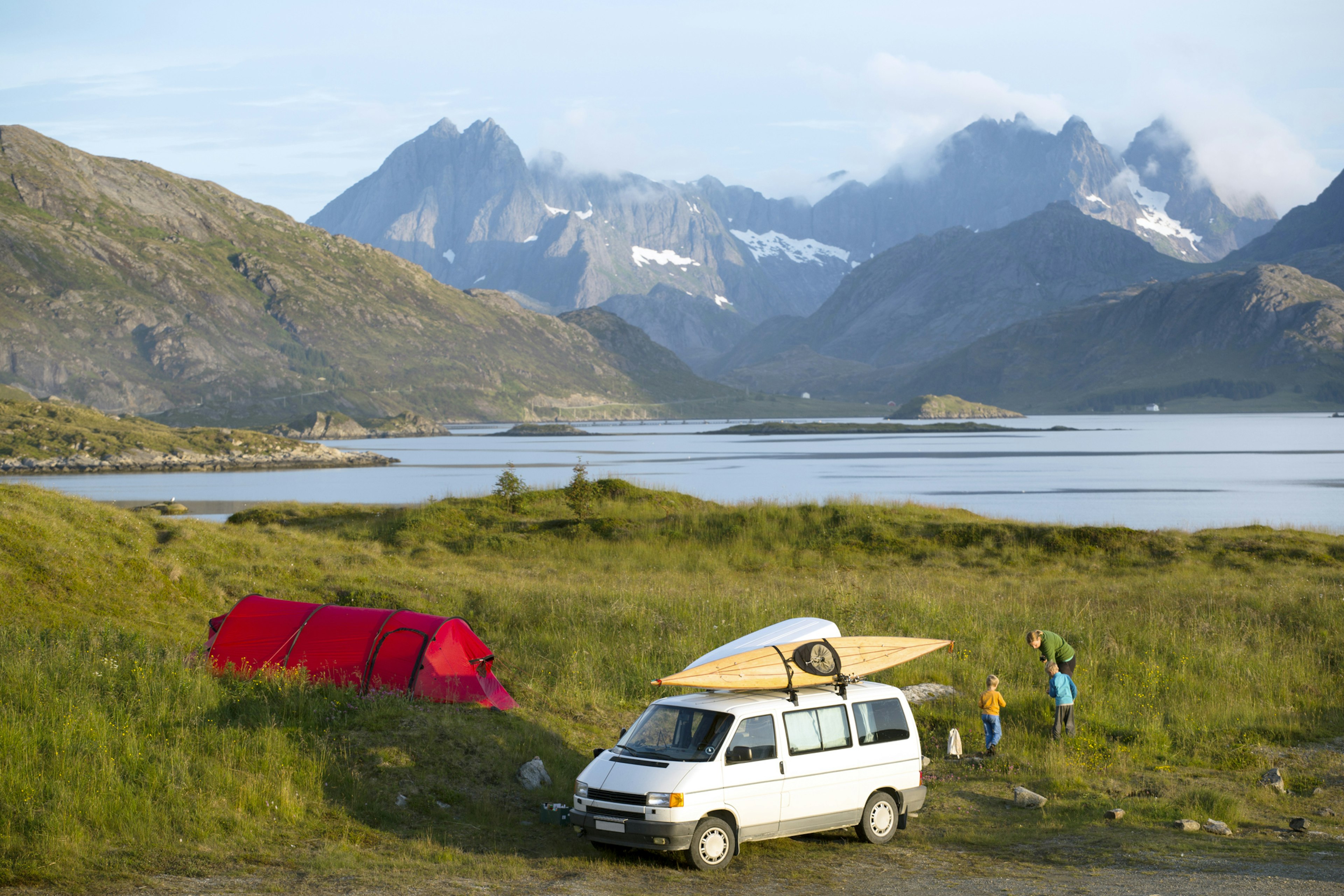 A mother and two children wander near a campervan parked by a lake in a mountainous area