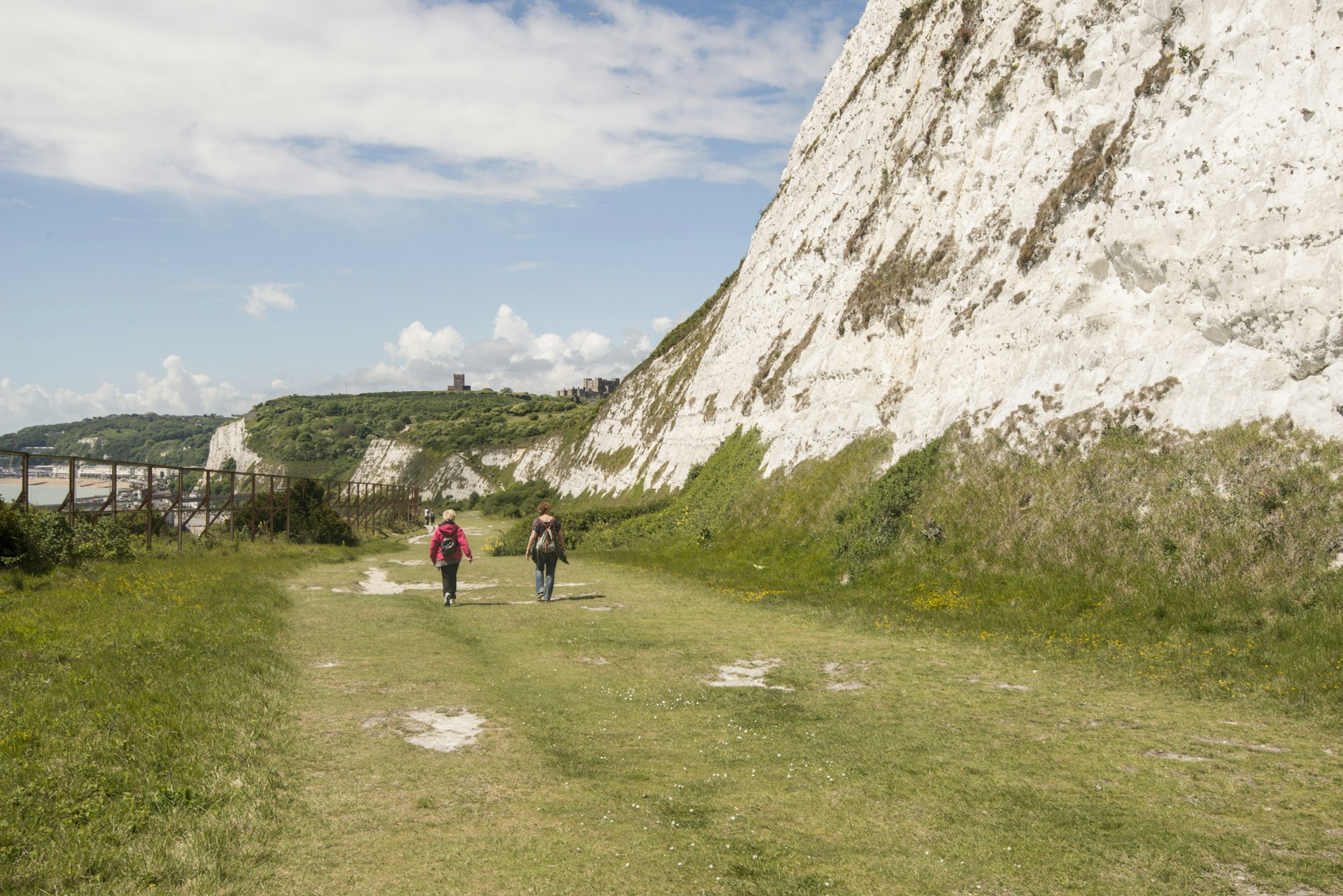 Two hikers walk on a path near the White Cliffs of Dover with a large castle on the clifftop in the distance