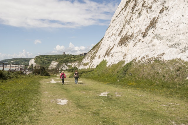 Two hikers walk on a path near the White Cliffs of Dover with a large castle on the clifftop in the distance