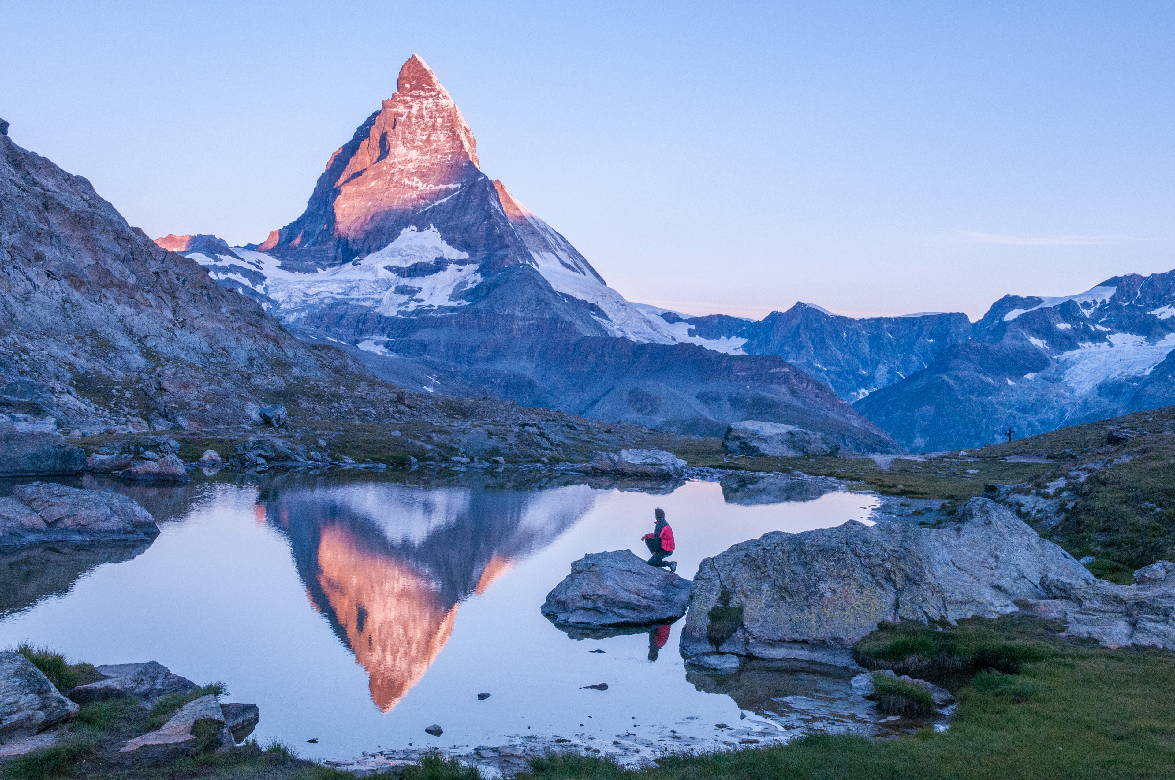 Early morning dawn scene of sunrise on the Matterhorn Mountain reflecting pink in the lake with male man on rock with red down jacket with clear blue sky, Zermatt