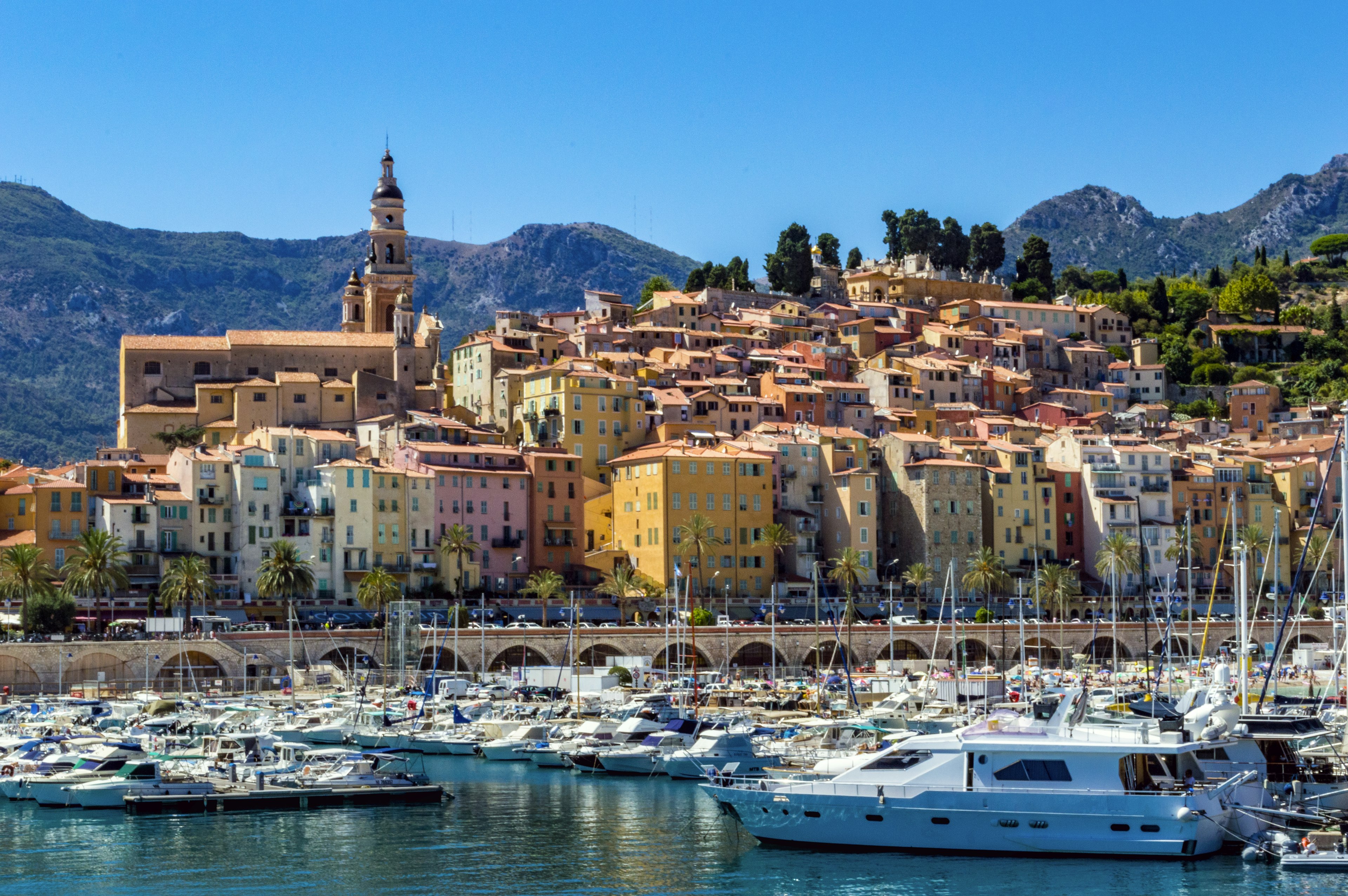 A huge church and buildings painted in pastel shades are built on a hill overlooking a marina filled with boats