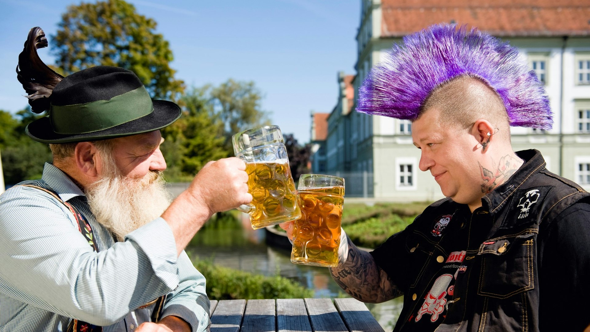 Two men, one in traditional Bavarian clothes, the other with a large purple mohawk, clink beer glasses 