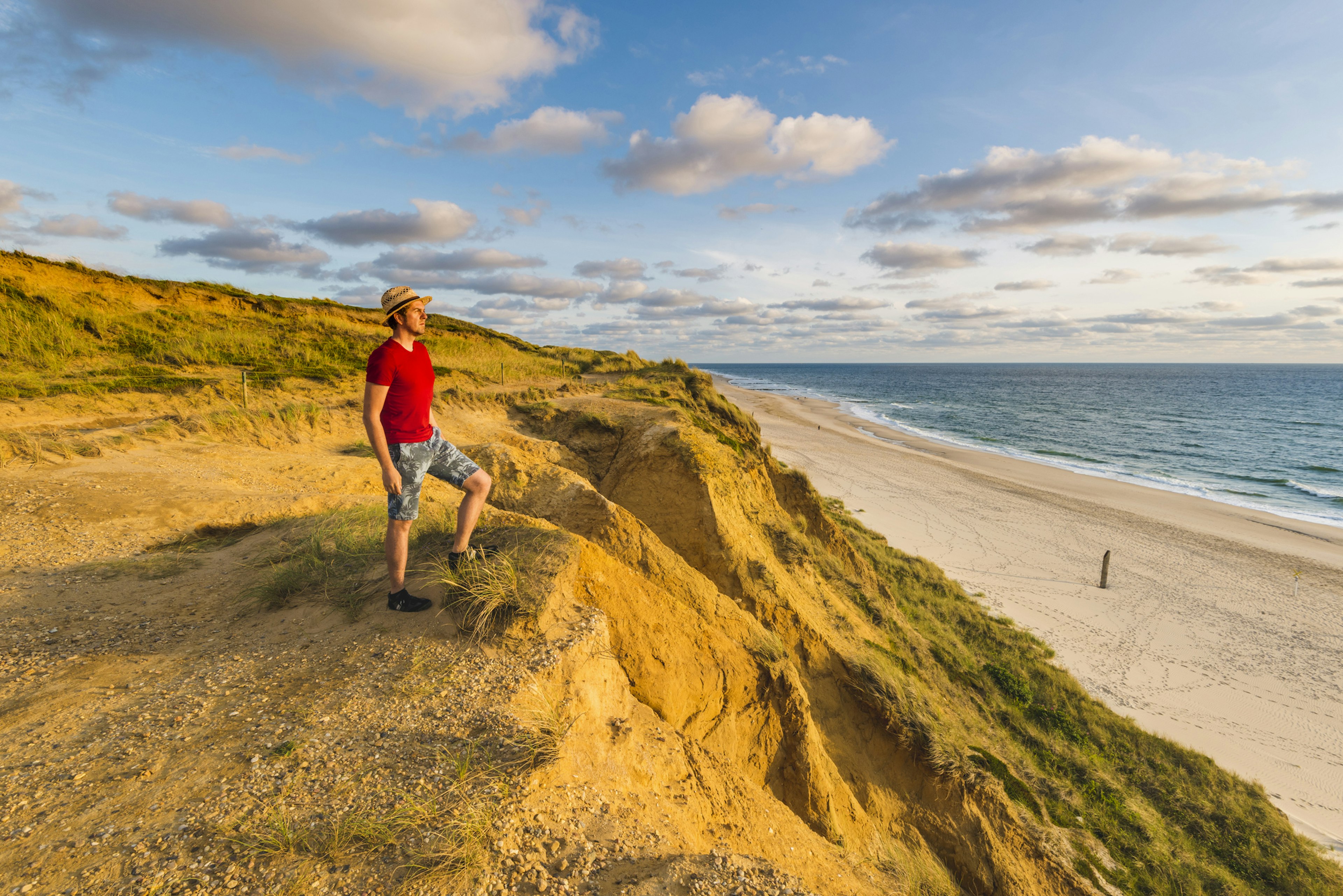 A man stands on a sandy dune and stares out over the beach towards the sea on a sunny day