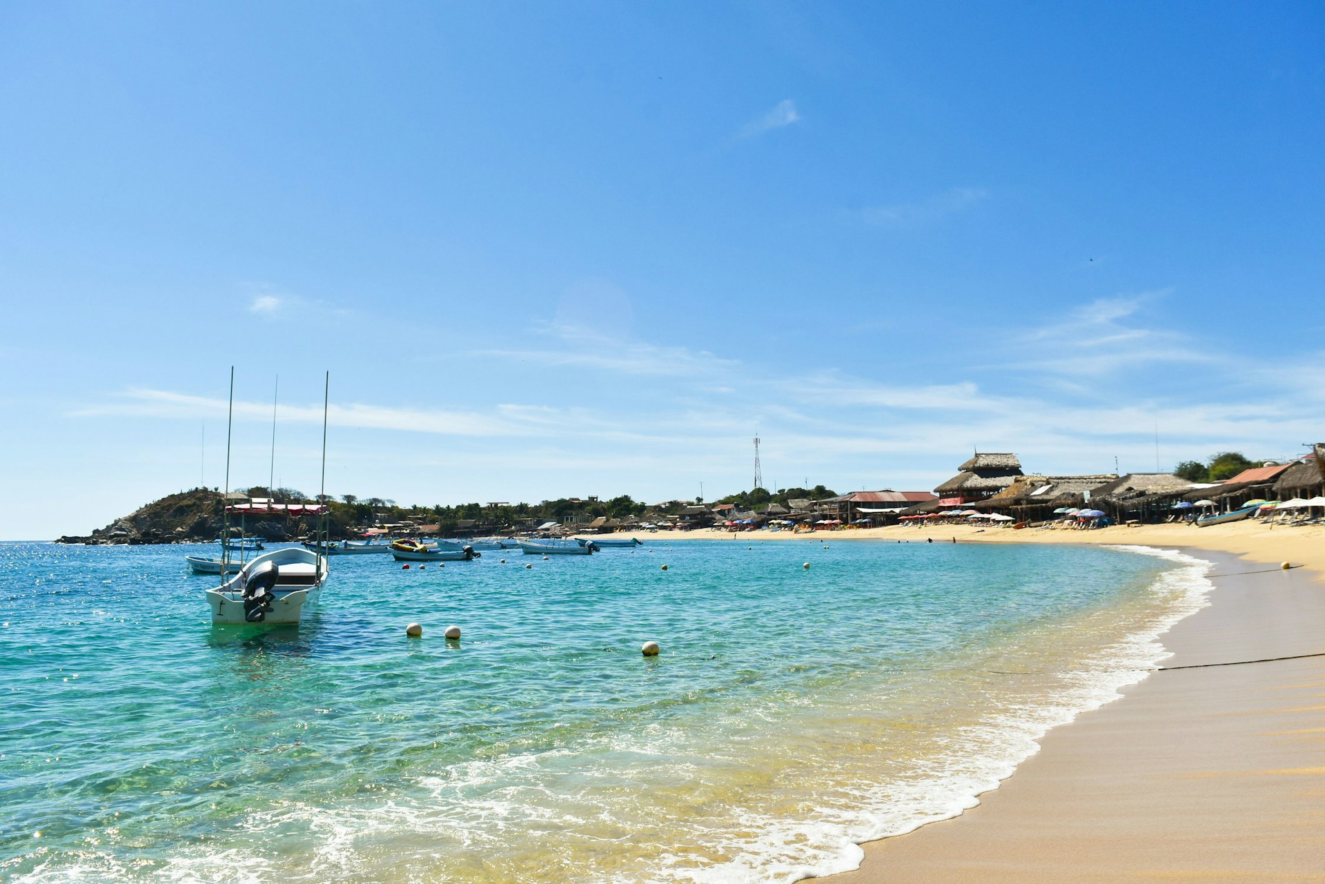 A picturesque beach scene under a clear blue sky. The sandy shore gently meets the calm turquoise waters, where a few small boats are anchored, bobbing with the waves.