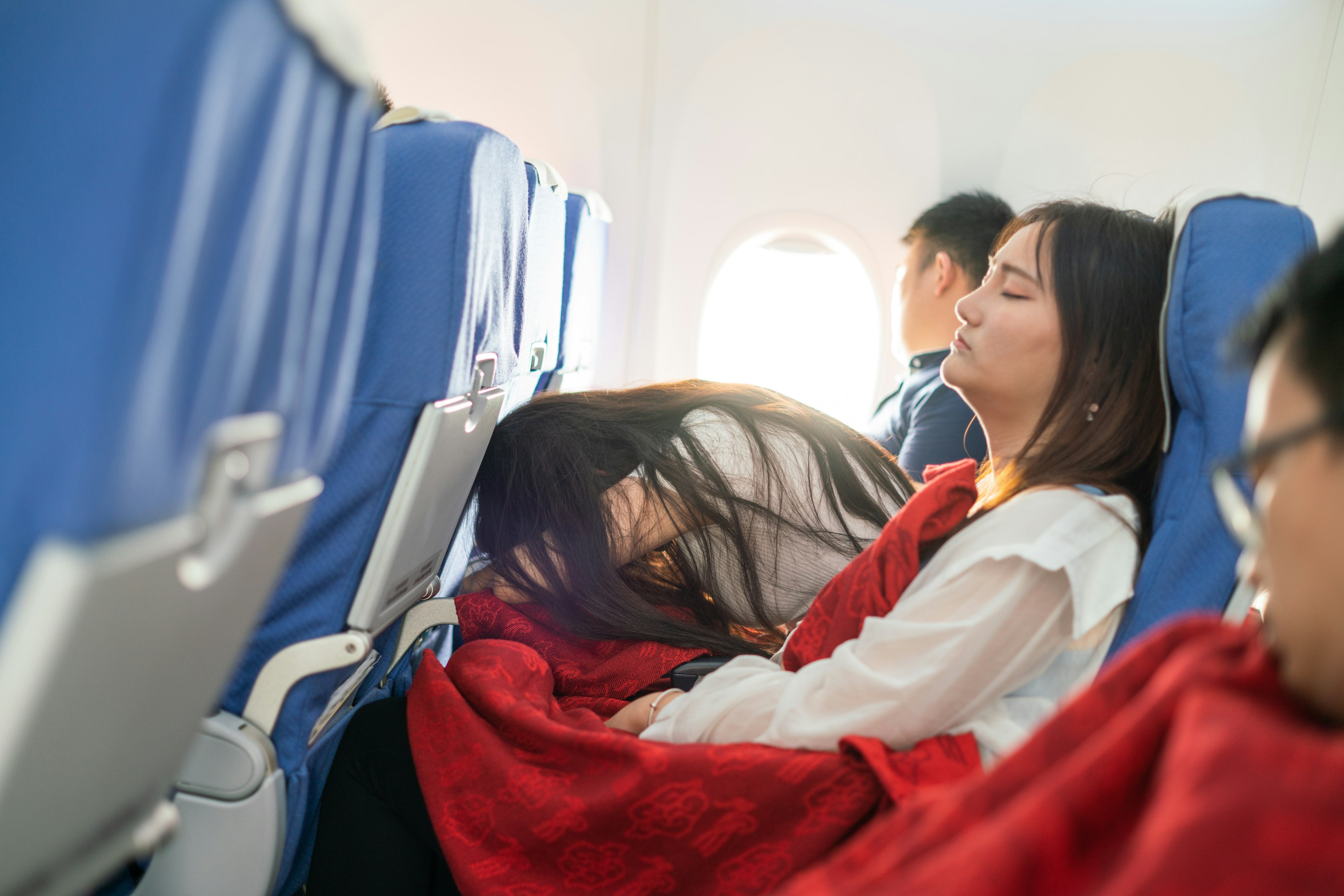 Inside the cabin of an airplane, a person in the middle seat is leaning over and sleeping on the tray-table.