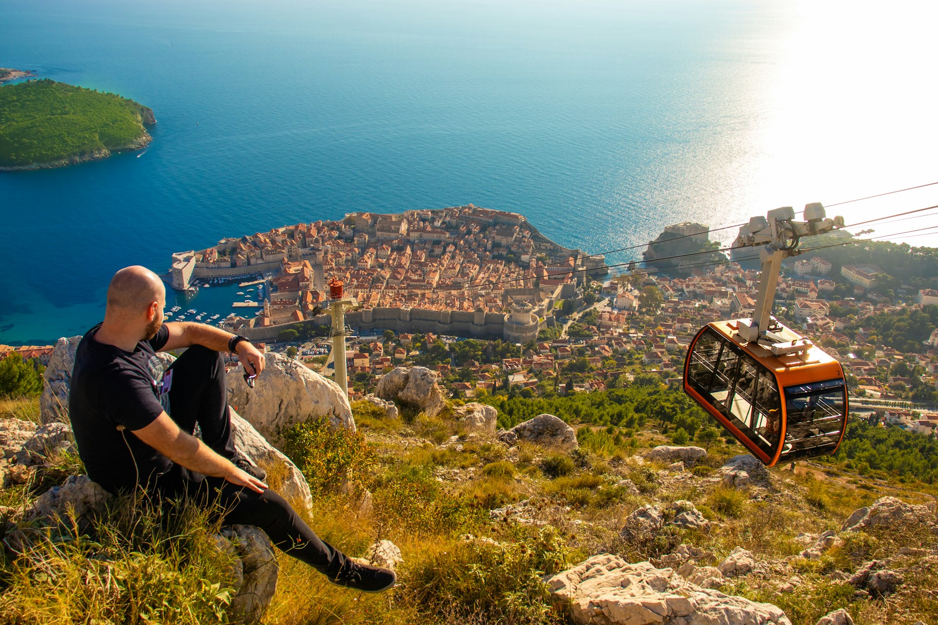 Wide shot from the top of the Srd mountain, man sitting on a rock observing the area around the city of Dubrovnik, orange cablecar descends to the city walls carrying tourist travelers in the summer.