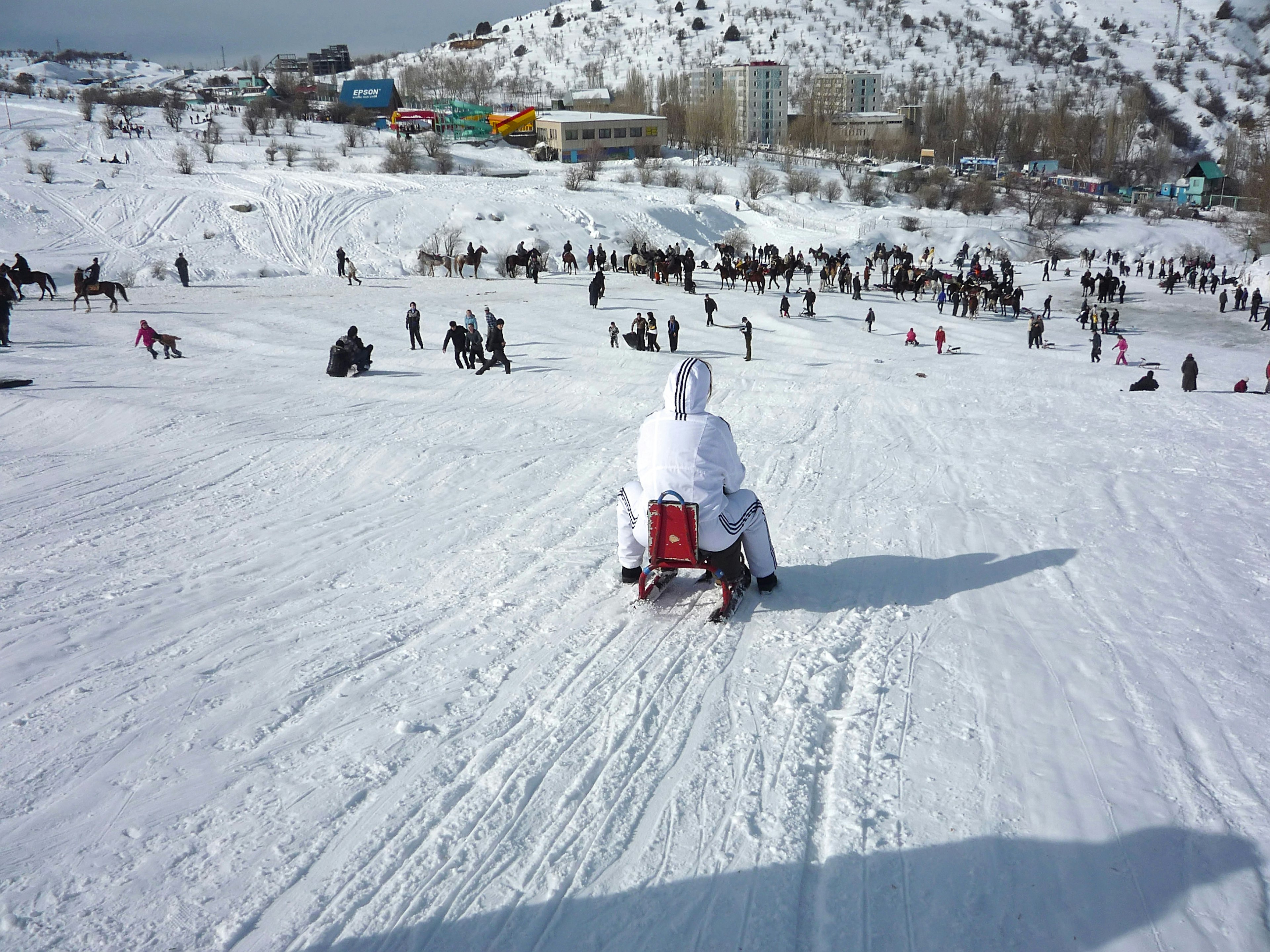 A child on a sledge heads down a slope dotted with people playing in the snow