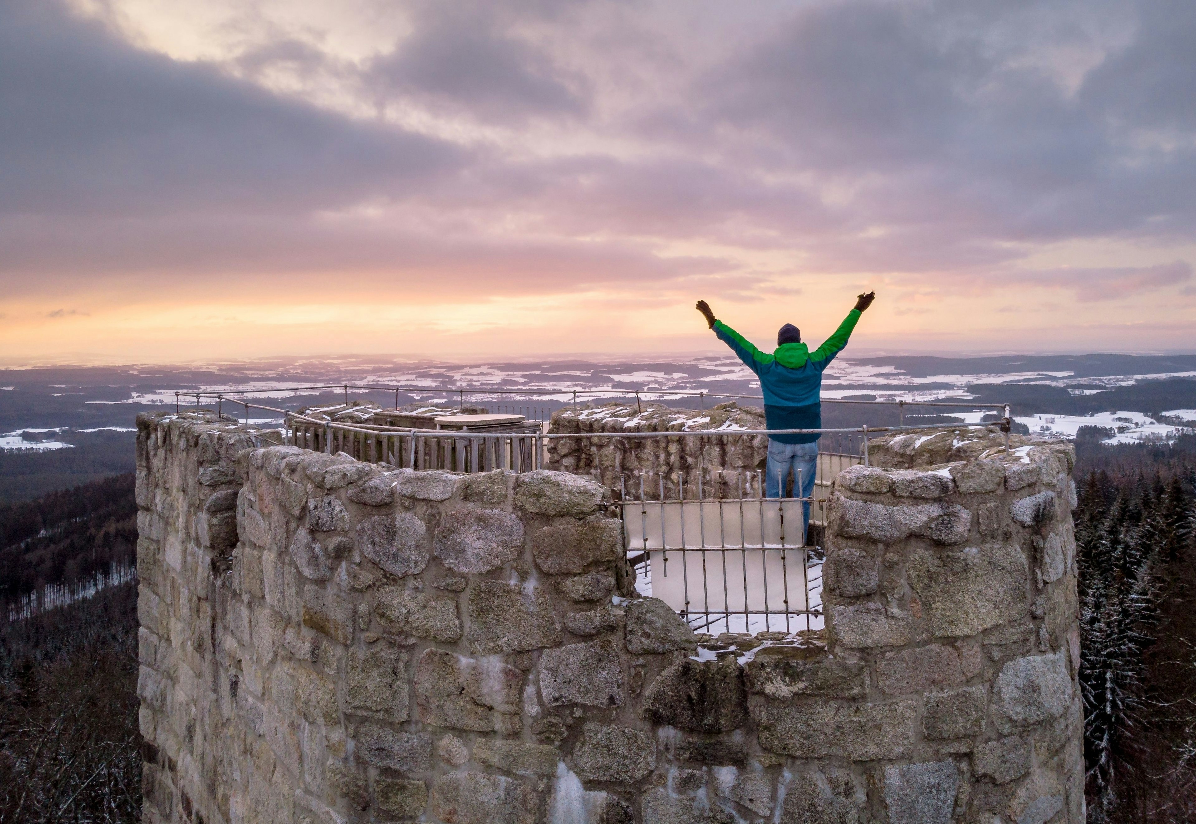 A man in winter gear stands on the top of a ruined castle tower looking out at the snowp-covered landscape