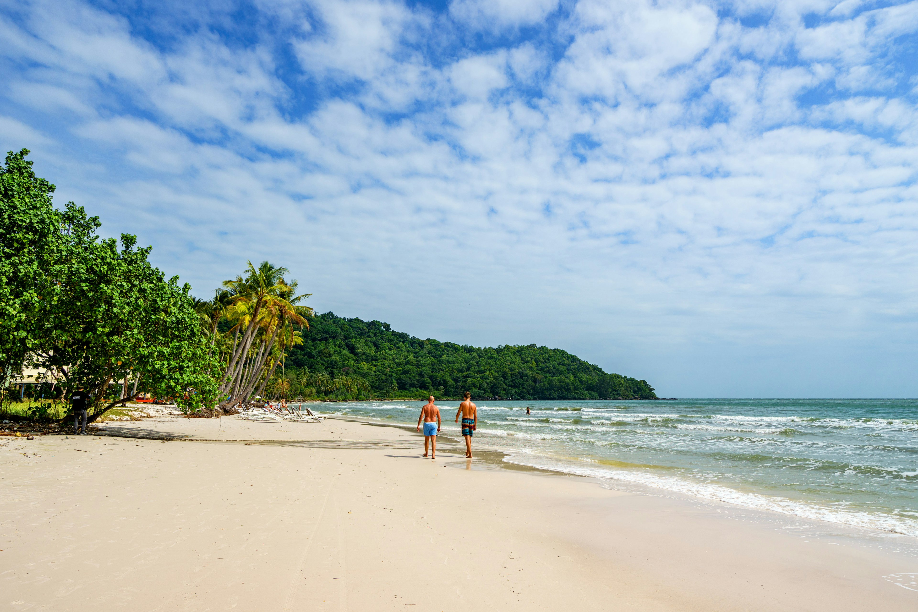 Two people in bathing suits walk on the white sandy Sao beach on Phy Quoc island, Vietnam. The ocean lapping the shore is blue and palm trees are visible.