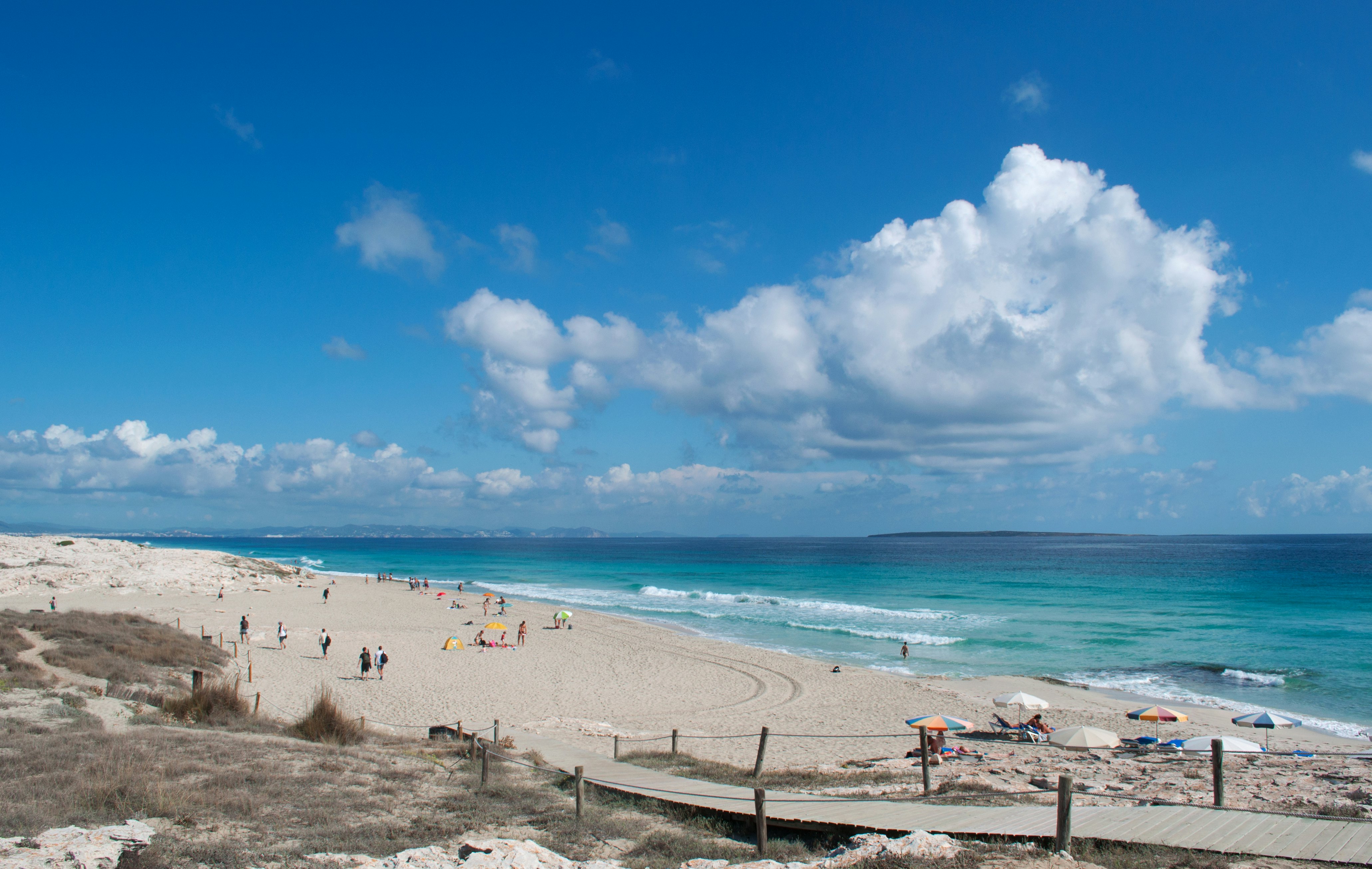 People under umbrellas relax on the sand of a large beach lapped by turquoise waves.