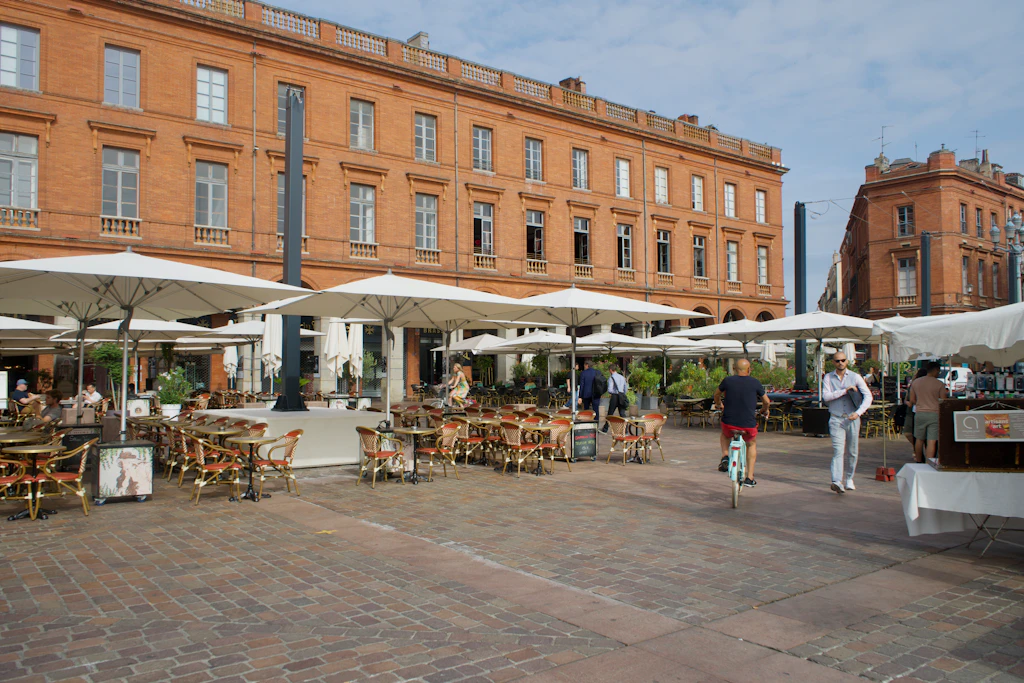 A series of restaurant tables with large white umbrellas line a square backed by a large red-brick building