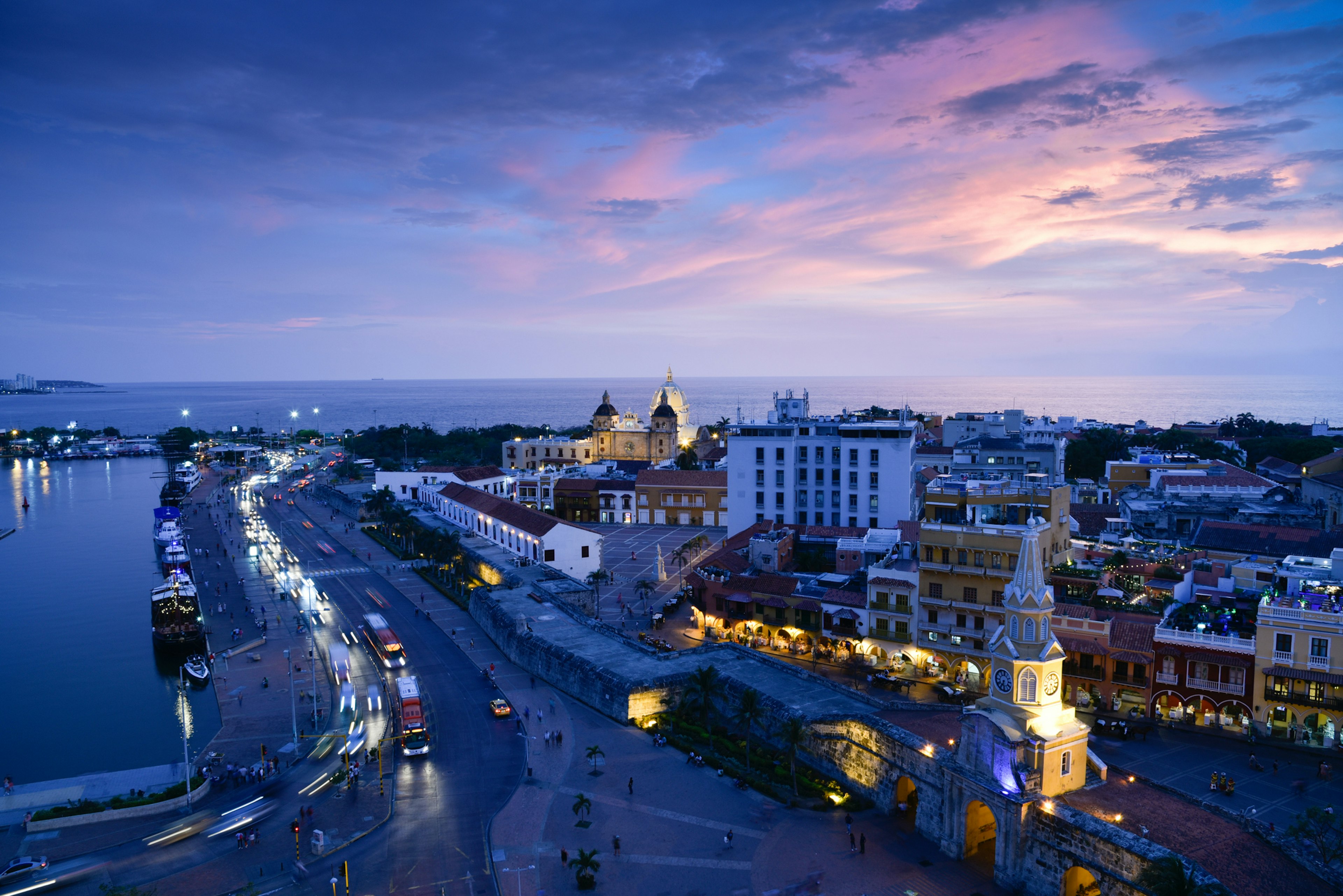 An aerial view of line of cars driving along the historic walls of Cartagena de Indias, with clock tower, at in the purple light of sunset, Colombia