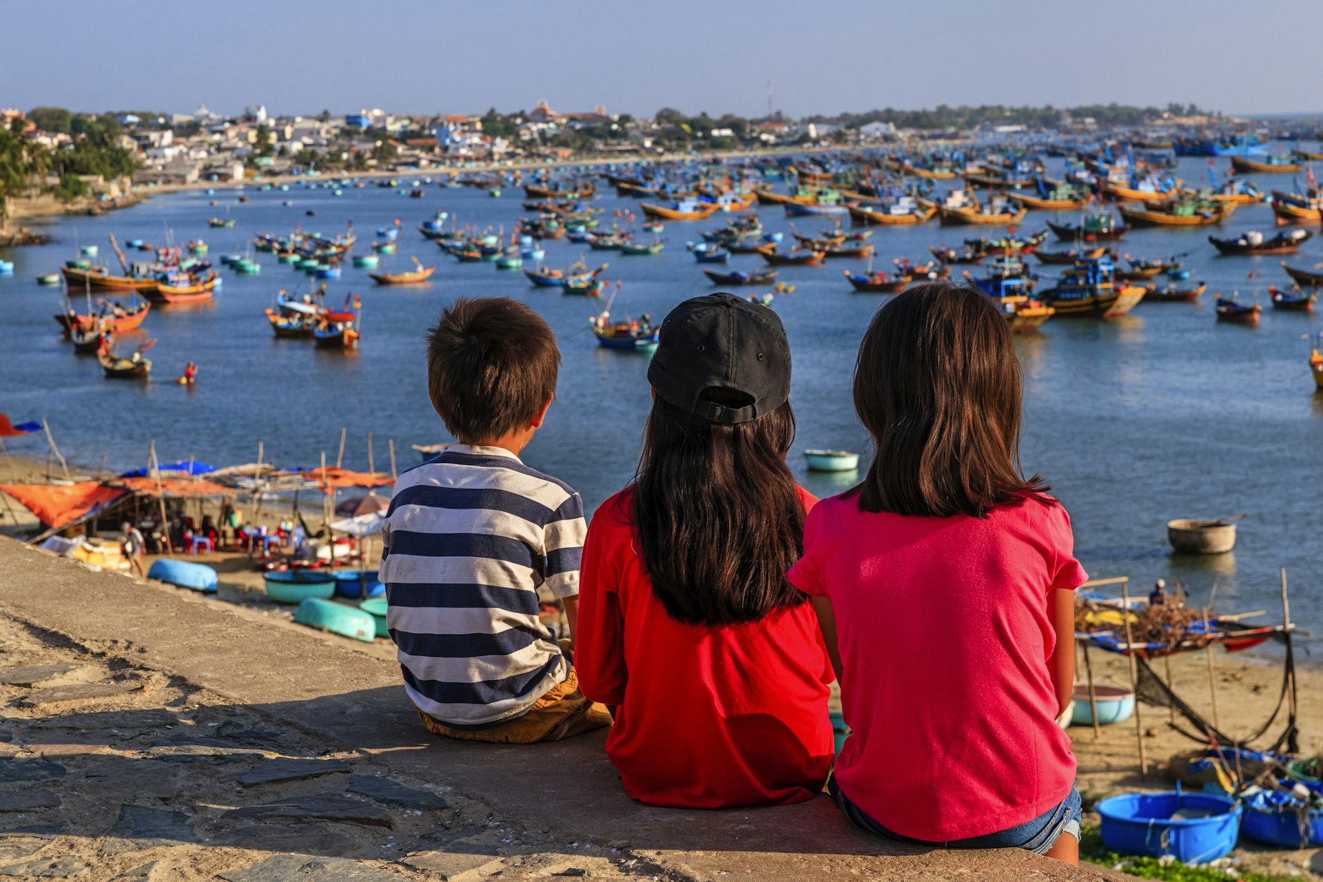 Three children sit on a wall looking out over a beach to the sea where many fishing boats have docked in the bay