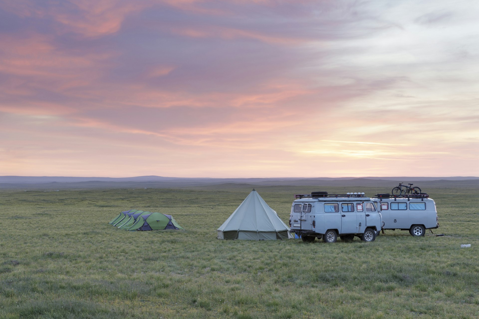 A green camping tent and a pointed white bell tent stand next to two four-wheel drive buses