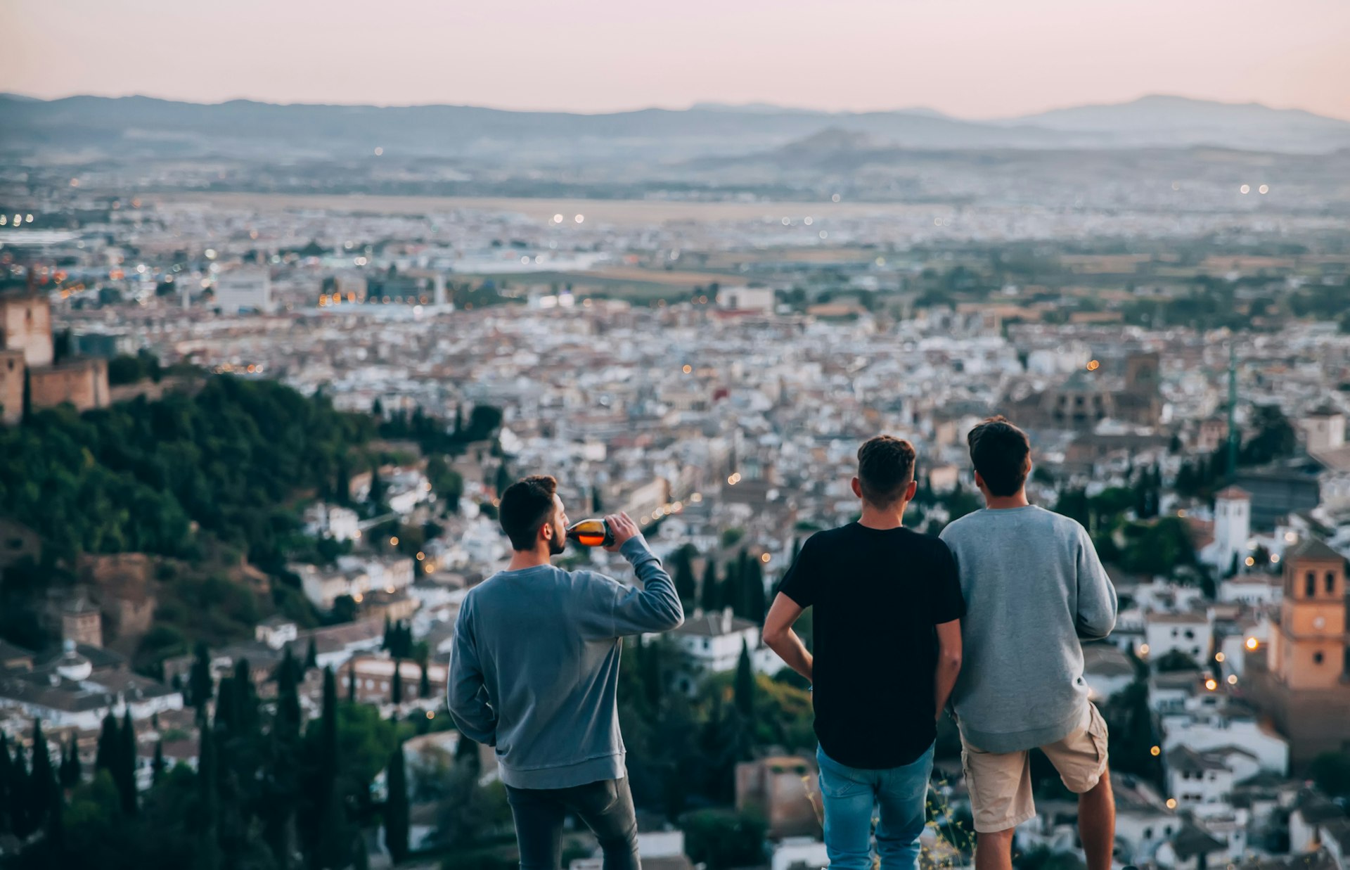 Three figures stand at a high-up viewpoint looking out across a city towards mountains