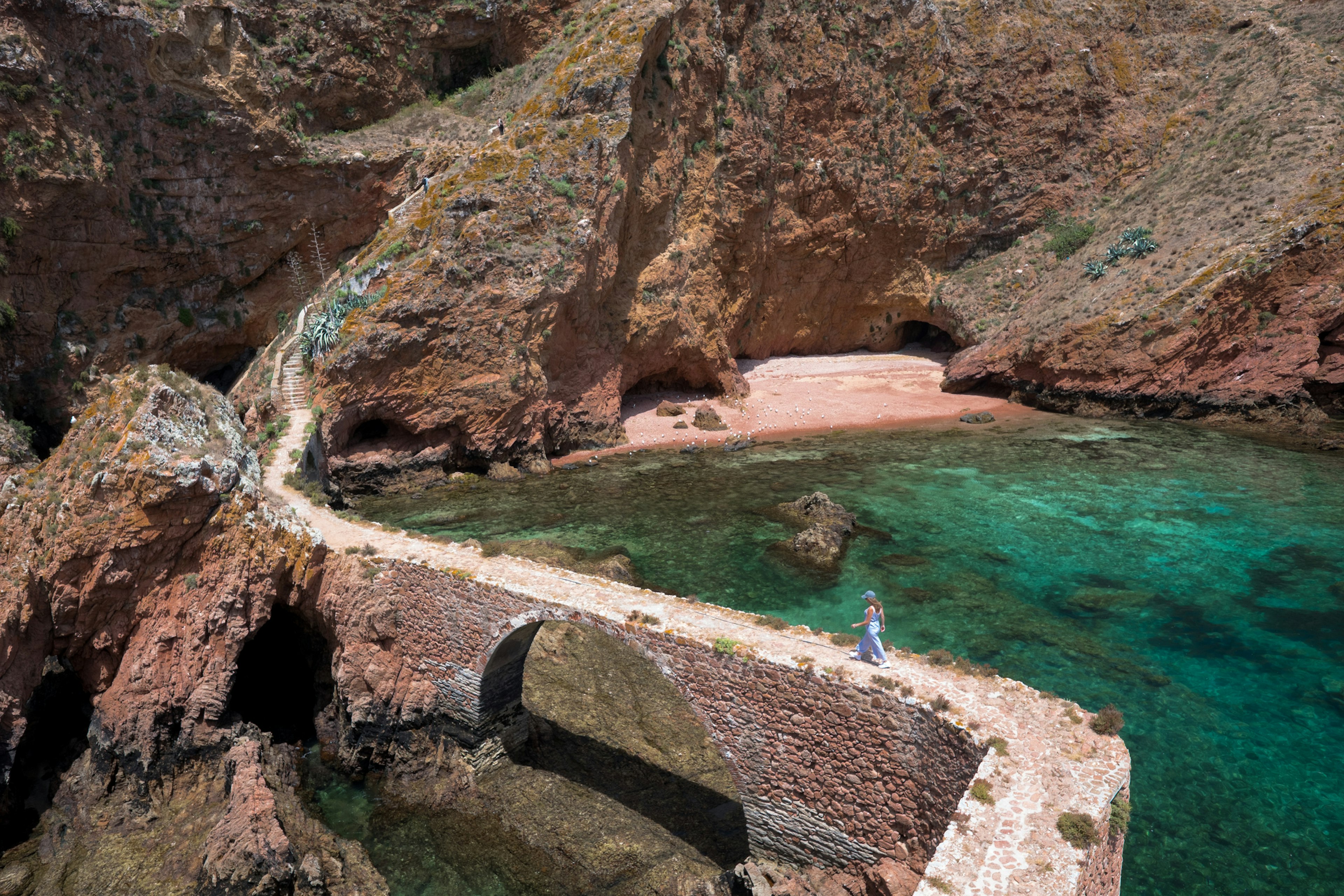 A figure follows a narrow stone bridge towards an island