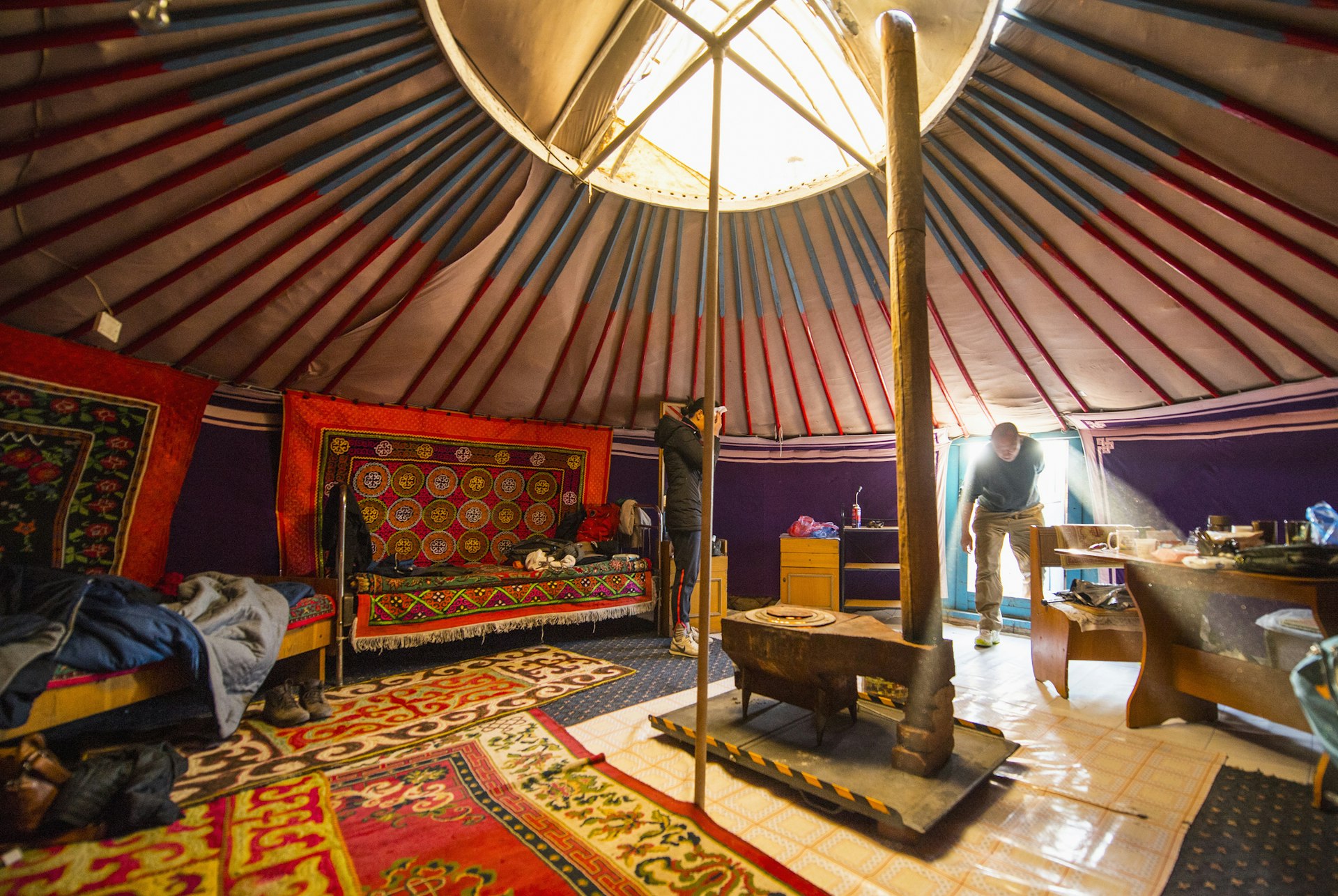 People step inside the doorway of a large round tent with a central opening and many colorful rugs adorning walls and floor