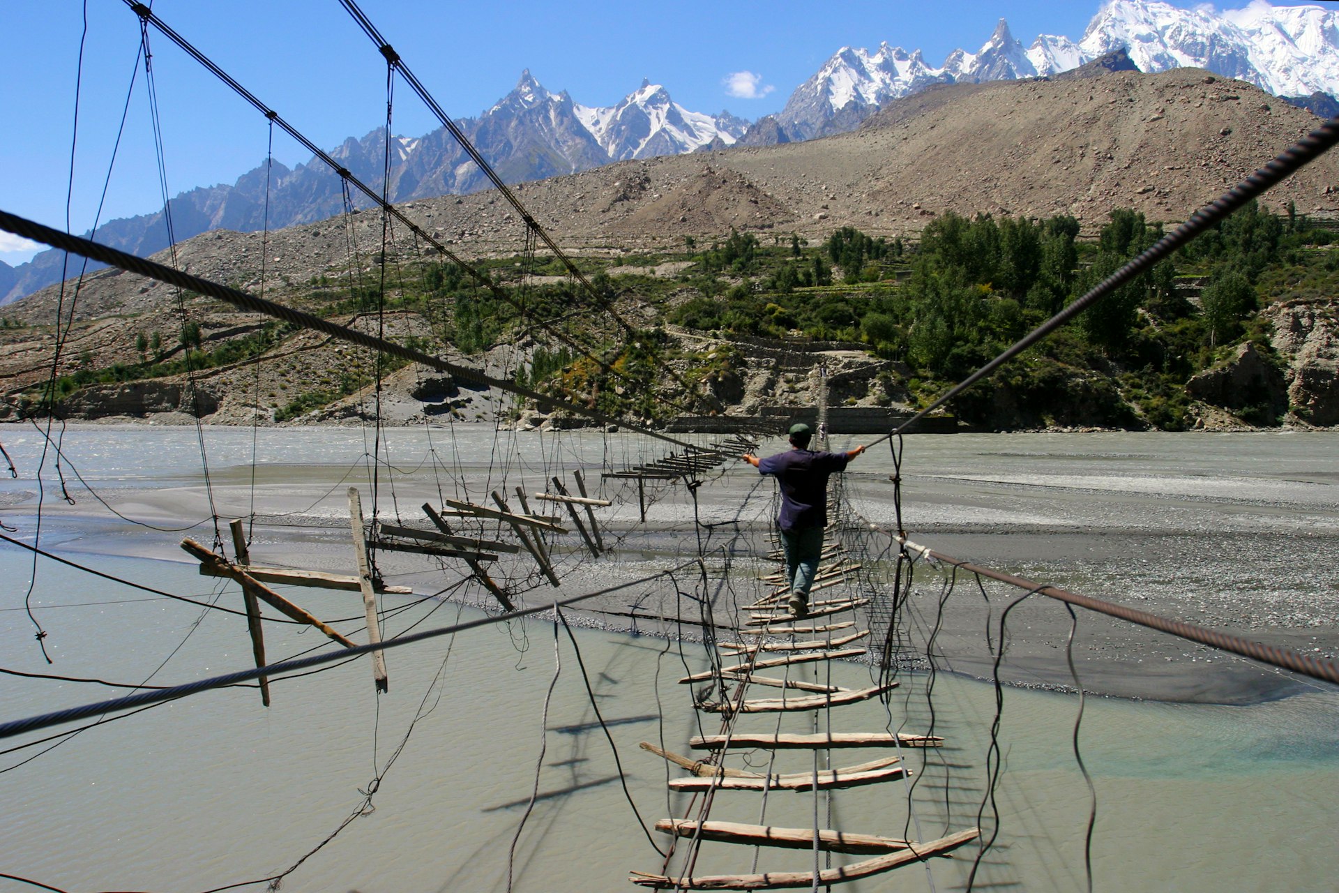 A man walks across a crude bridge strung together with ropes and wooden slats over a river 