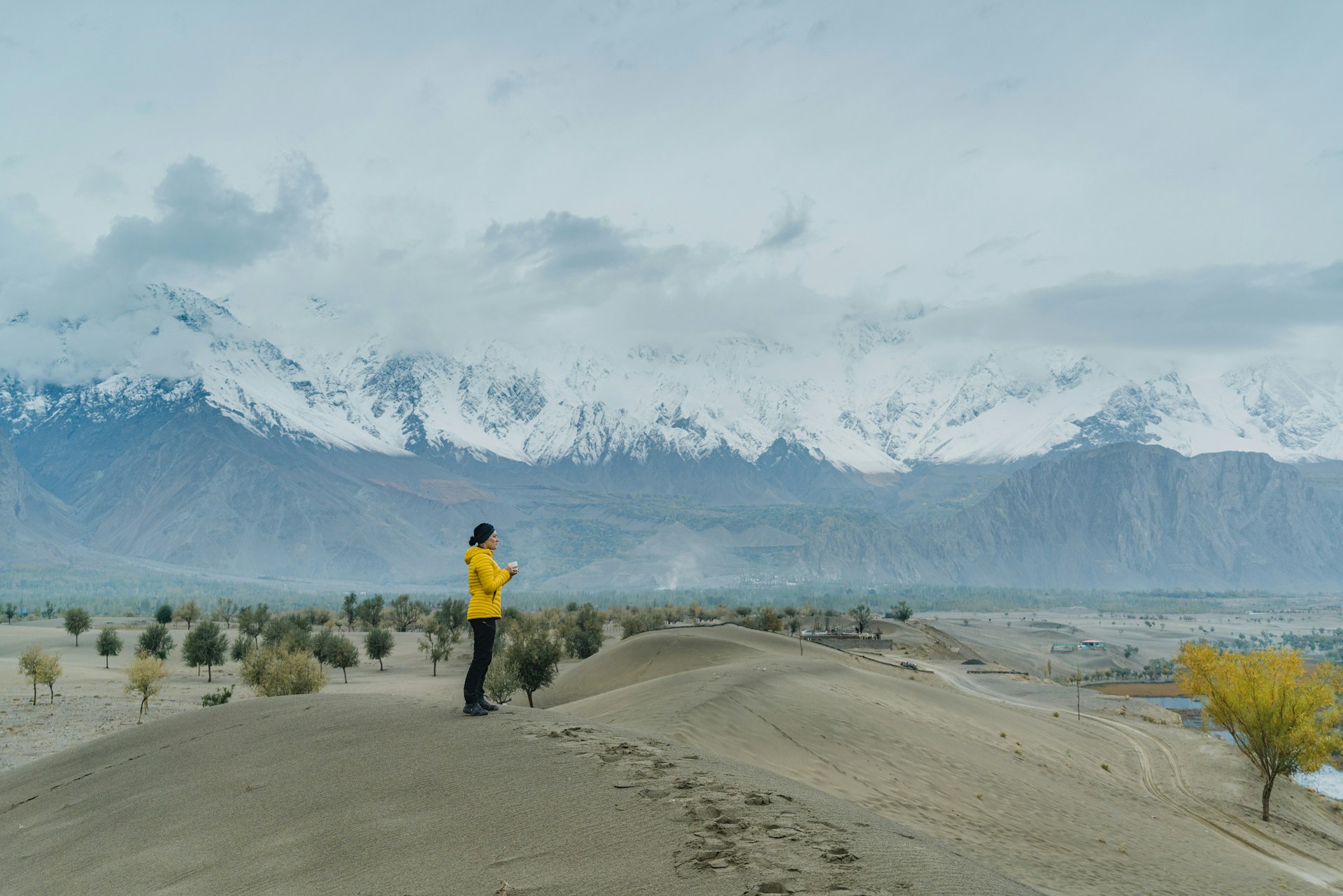 A woman stands on a sandy plain with a range of snowcapped mountains rising in the distance