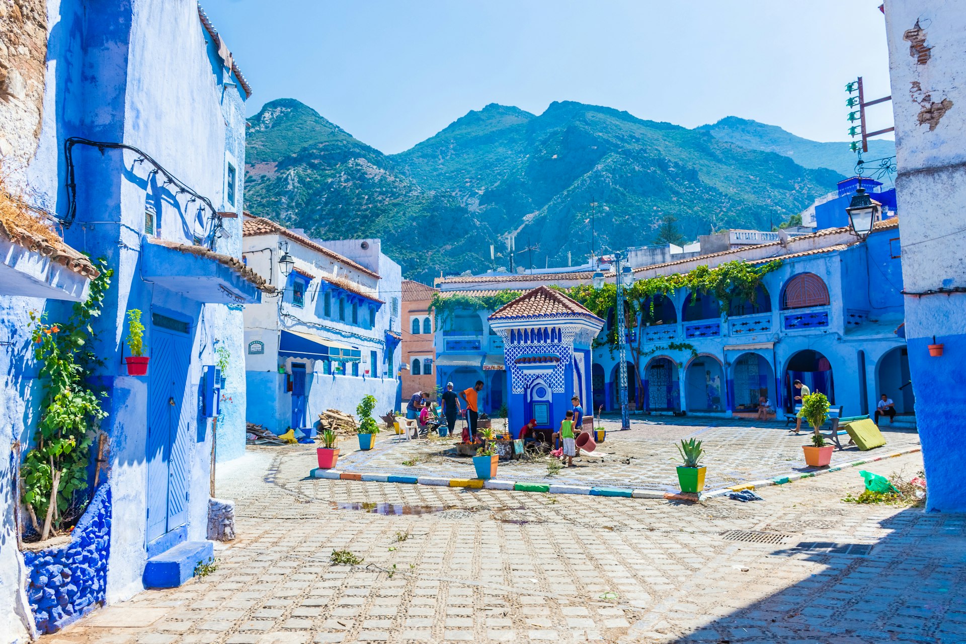 A small cobbled square surrounded by low-rise buildings that are painted blue. Mountain peaks rise in the distance