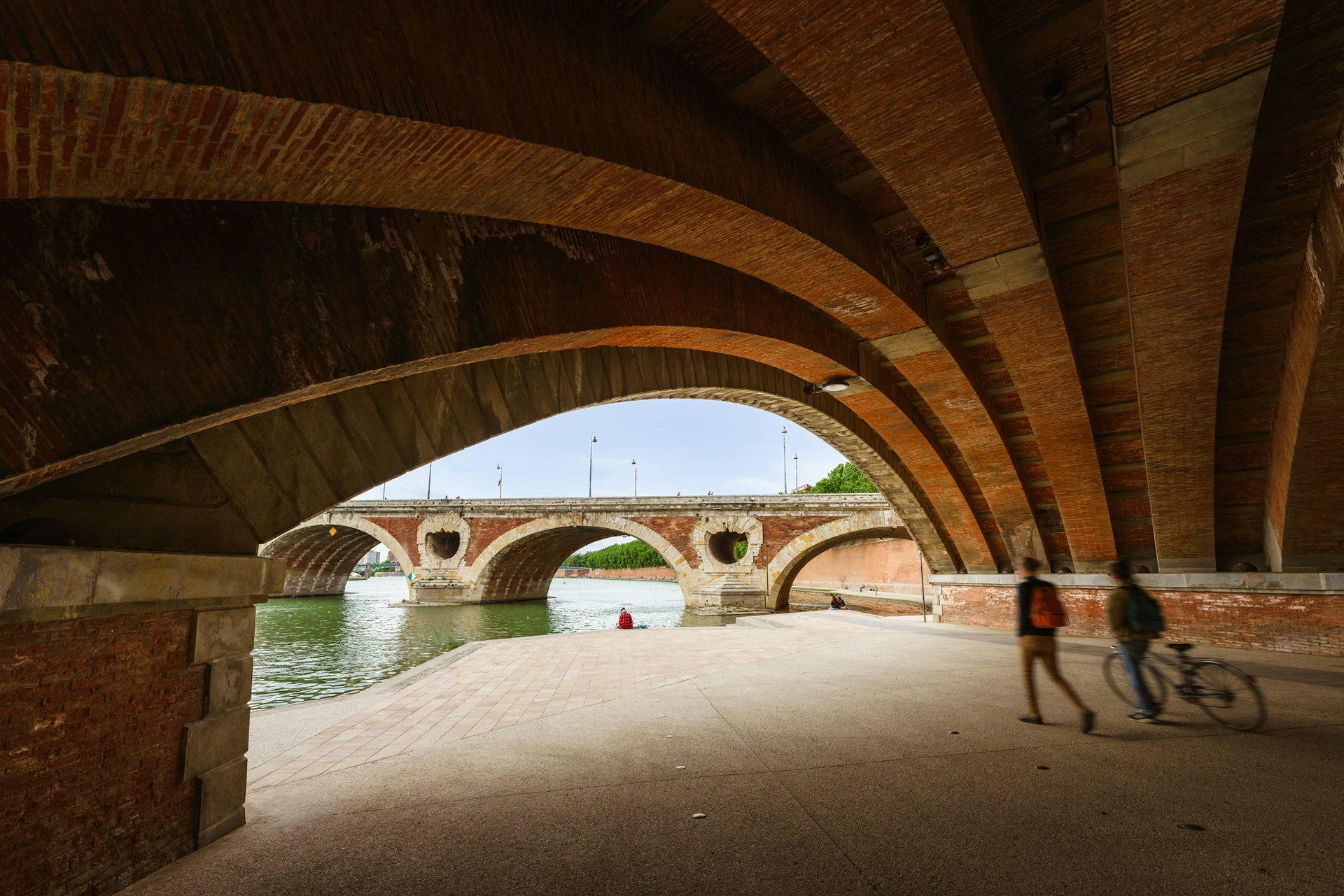 Two people wheel a bike through a pedestrianized walkway under a bridge to a path alongside a river