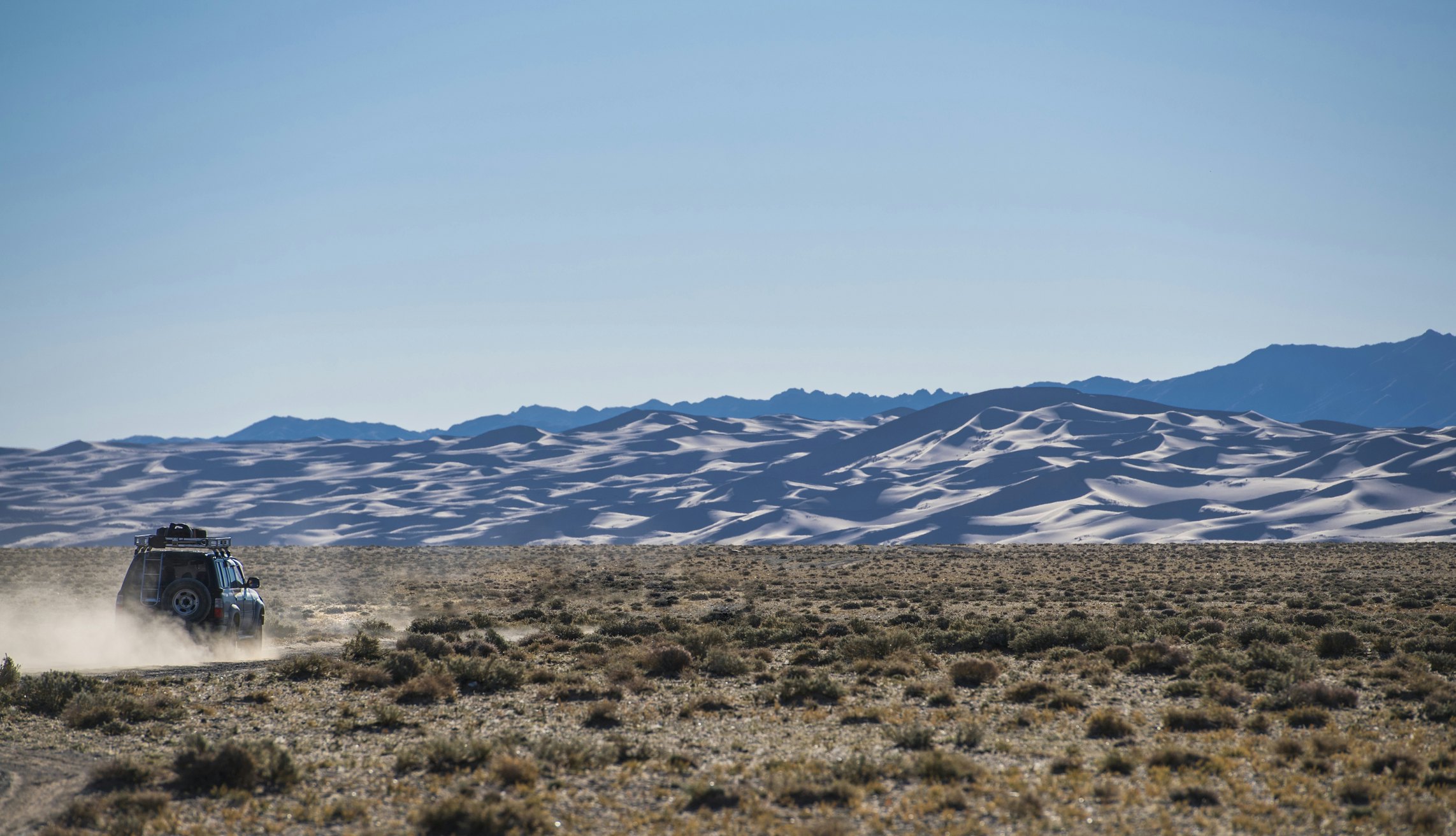 A four-wheel-drive vehicle drives a dusty track towards sand dunes in a desert