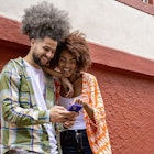 Latin American female friends between 25 and 30 years old looking at the cell phone leaning against the wall of the red wall while looking at social networks outdoors on the city street, she points with her index finger what she sees on the cell phone screen, the girl is African American with wavy brown hair and the young man has wavy hair with white skin.
1523005500
A man and woman leaning against a wall in Bogotá while smiling and looking at a smartphone