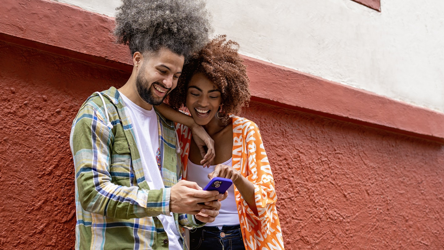 Latin American female friends between 25 and 30 years old looking at the cell phone leaning against the wall of the red wall while looking at social networks outdoors on the city street, she points with her index finger what she sees on the cell phone screen, the girl is African American with wavy brown hair and the young man has wavy hair with white skin.
1523005500
A man and woman leaning against a wall in Bogotá while smiling and looking at a smartphone