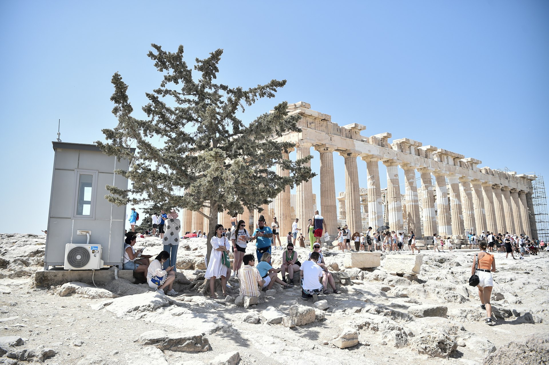 Atop the Acropolis ancient hill with Parthenon temple in background, tourists shelter from the sun under a tree 