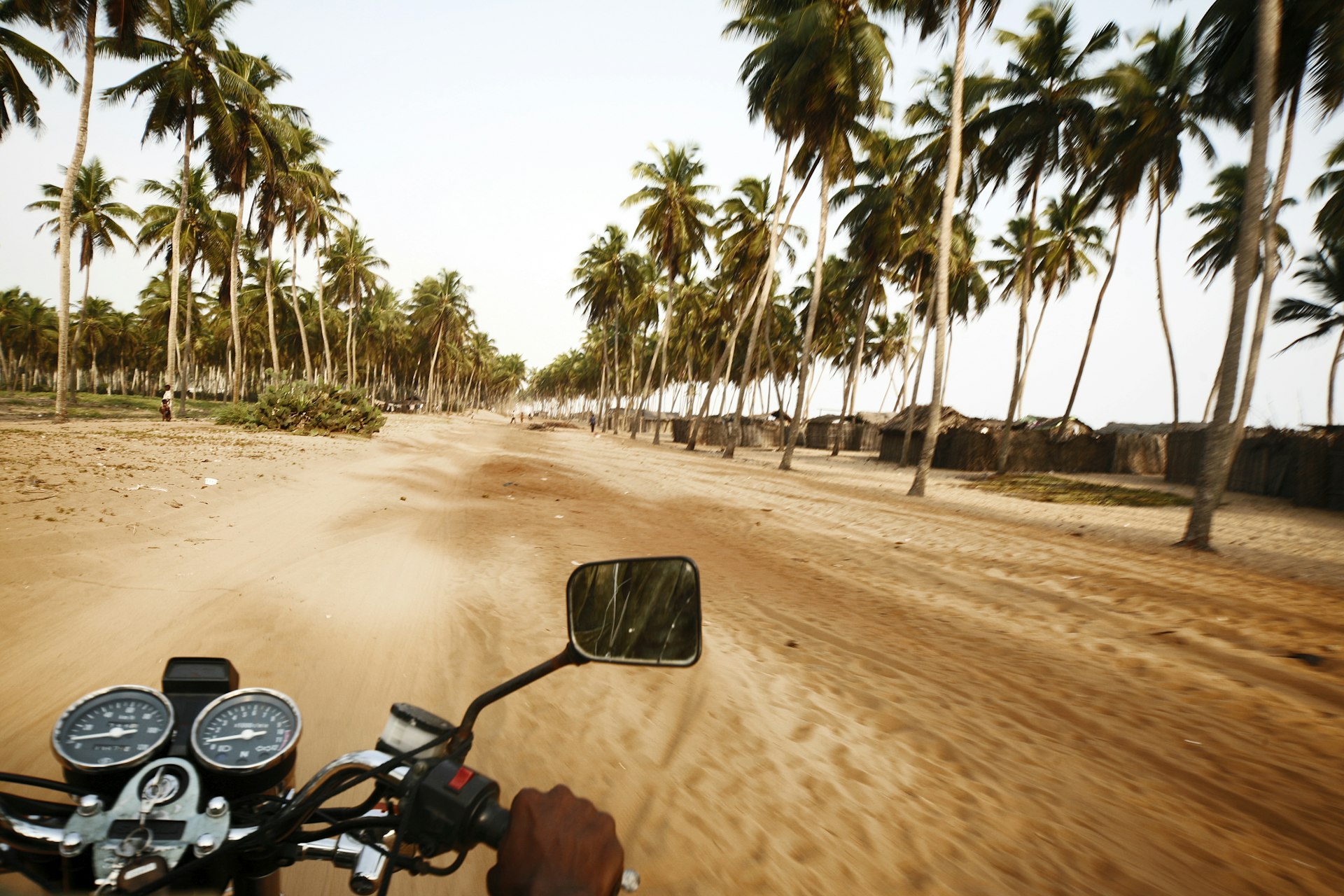 A motorcycle rides along a sandy palm-lined track alongside a beach