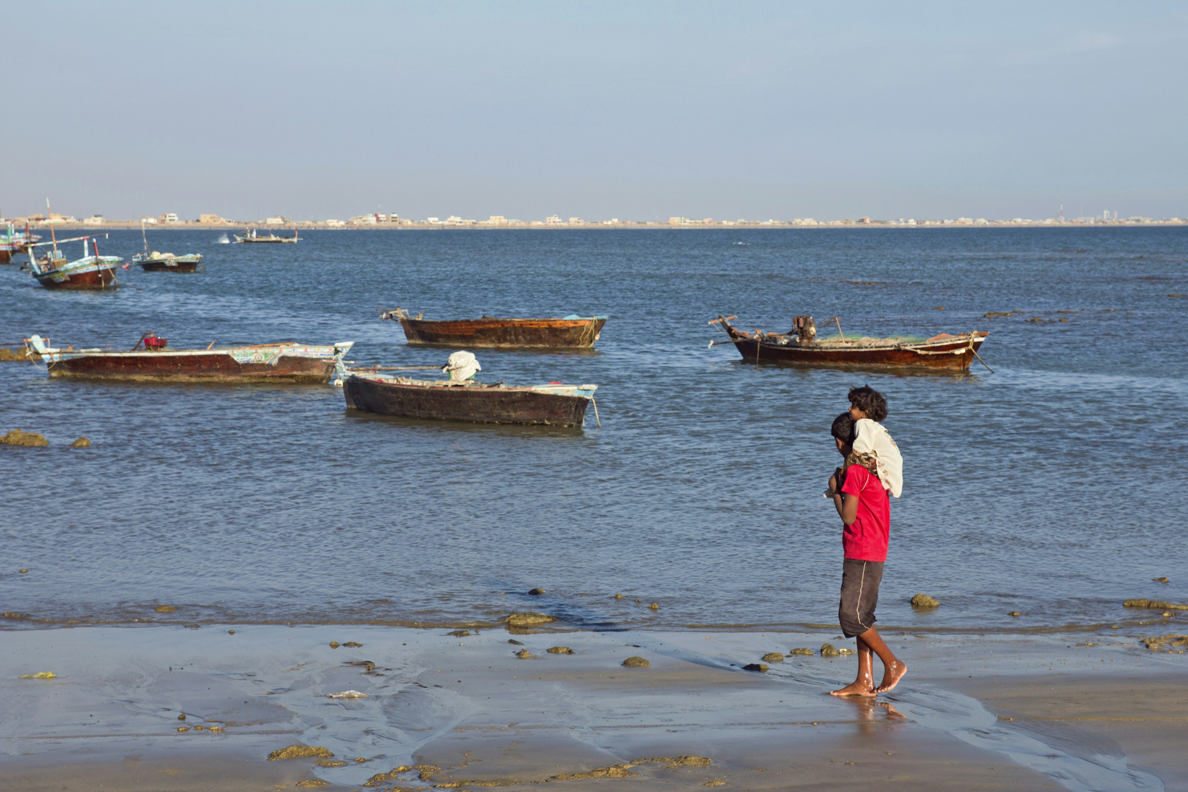 A girl is riding on her brother's shoulder along the water's edge by the sea