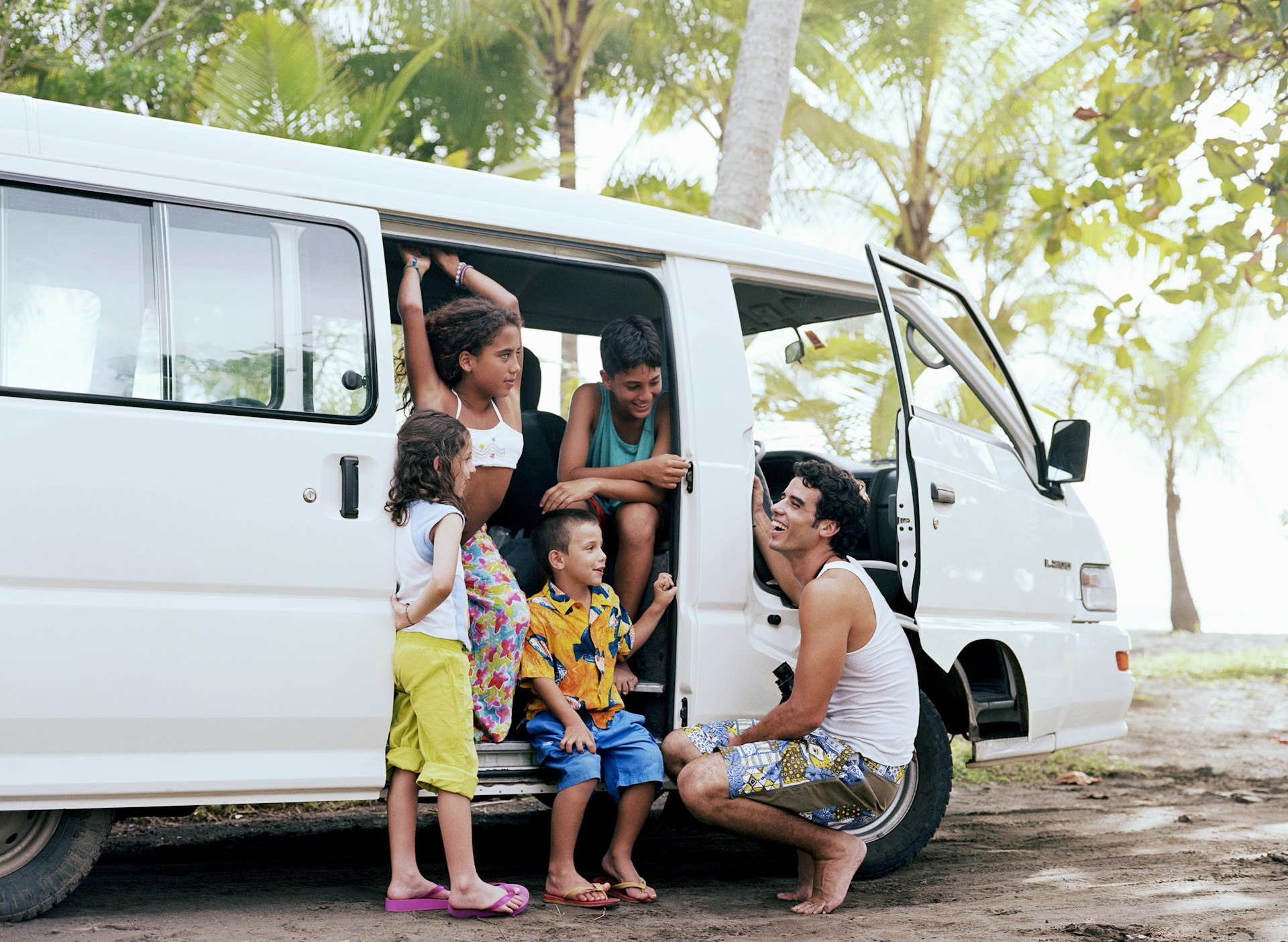 A man crouches down by the open door of a white camper van; four children are inside