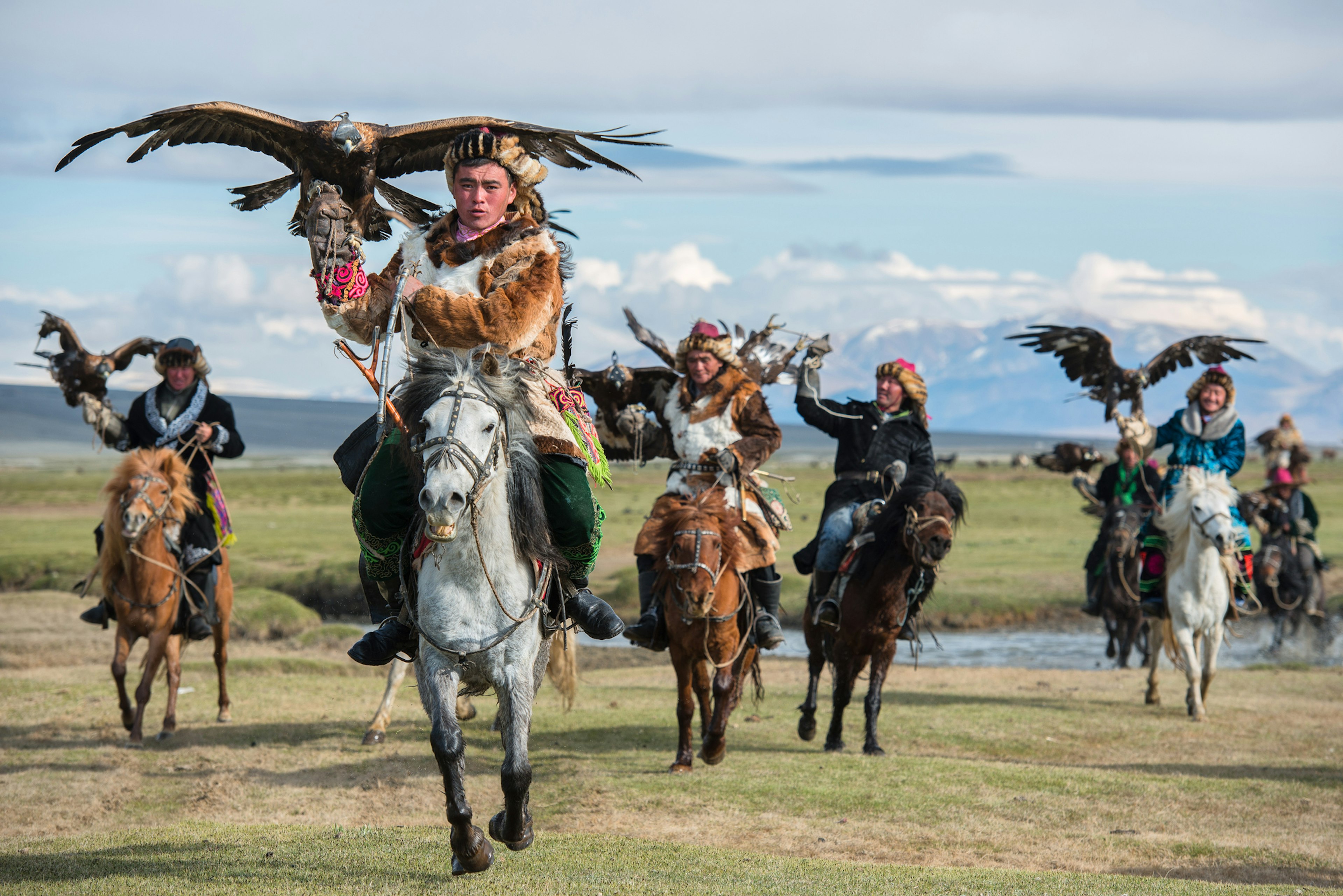 Horseback-riders holding eagles aloft with their wings spread out charge across a river and onto the grassy plains