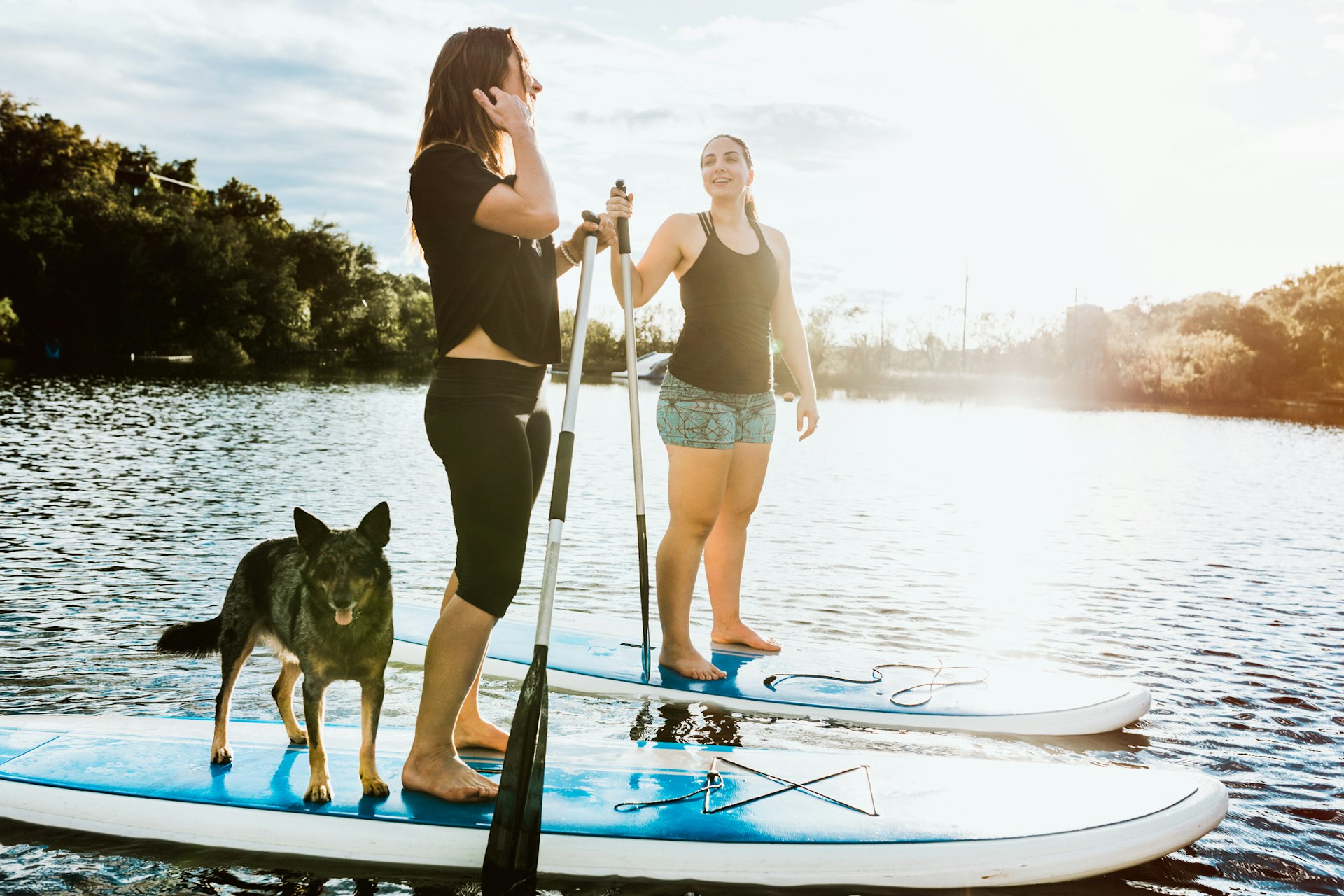 Two young women friends enjoys a peaceful moment on the water with paddle boards and a pet dog.  The sun illuminates the scene, casting a golden glow, in Austin, Texas