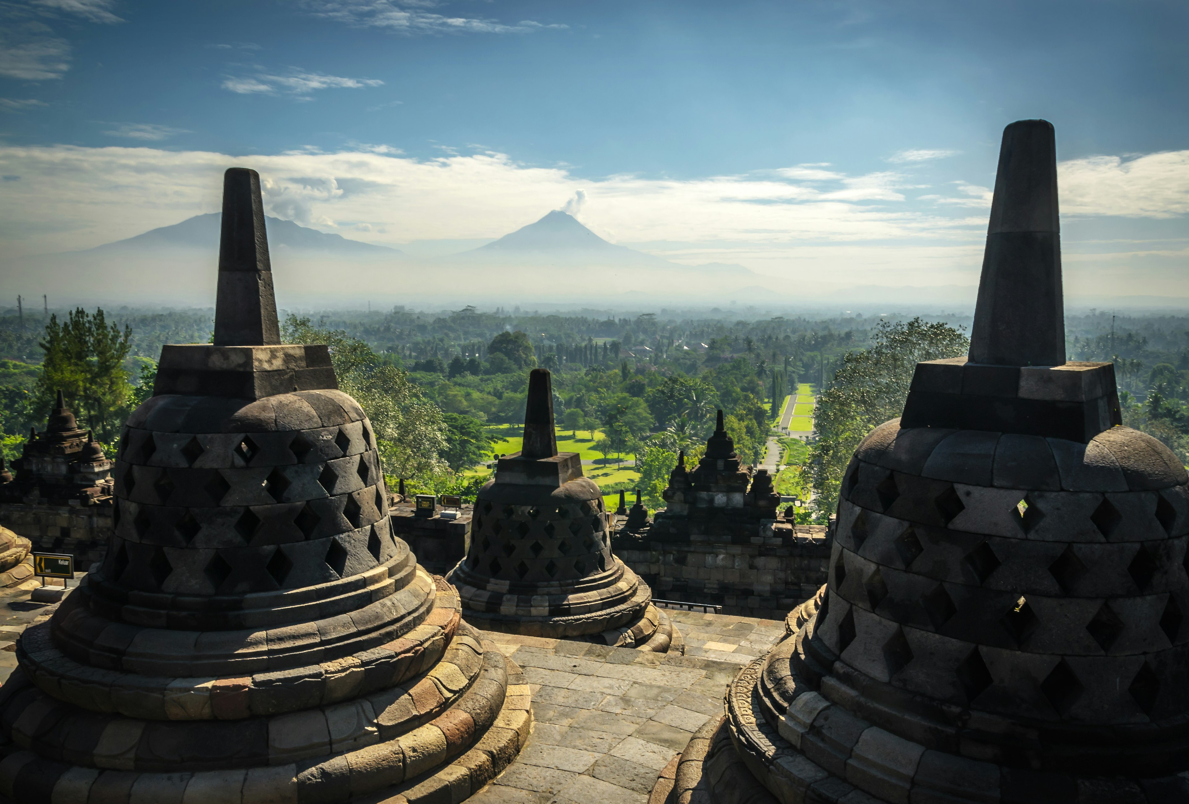 Bell-shaped stupa monuments on the upper level of a Buddhist temple stand above a green plain with distant peaks in Java, Indonesia