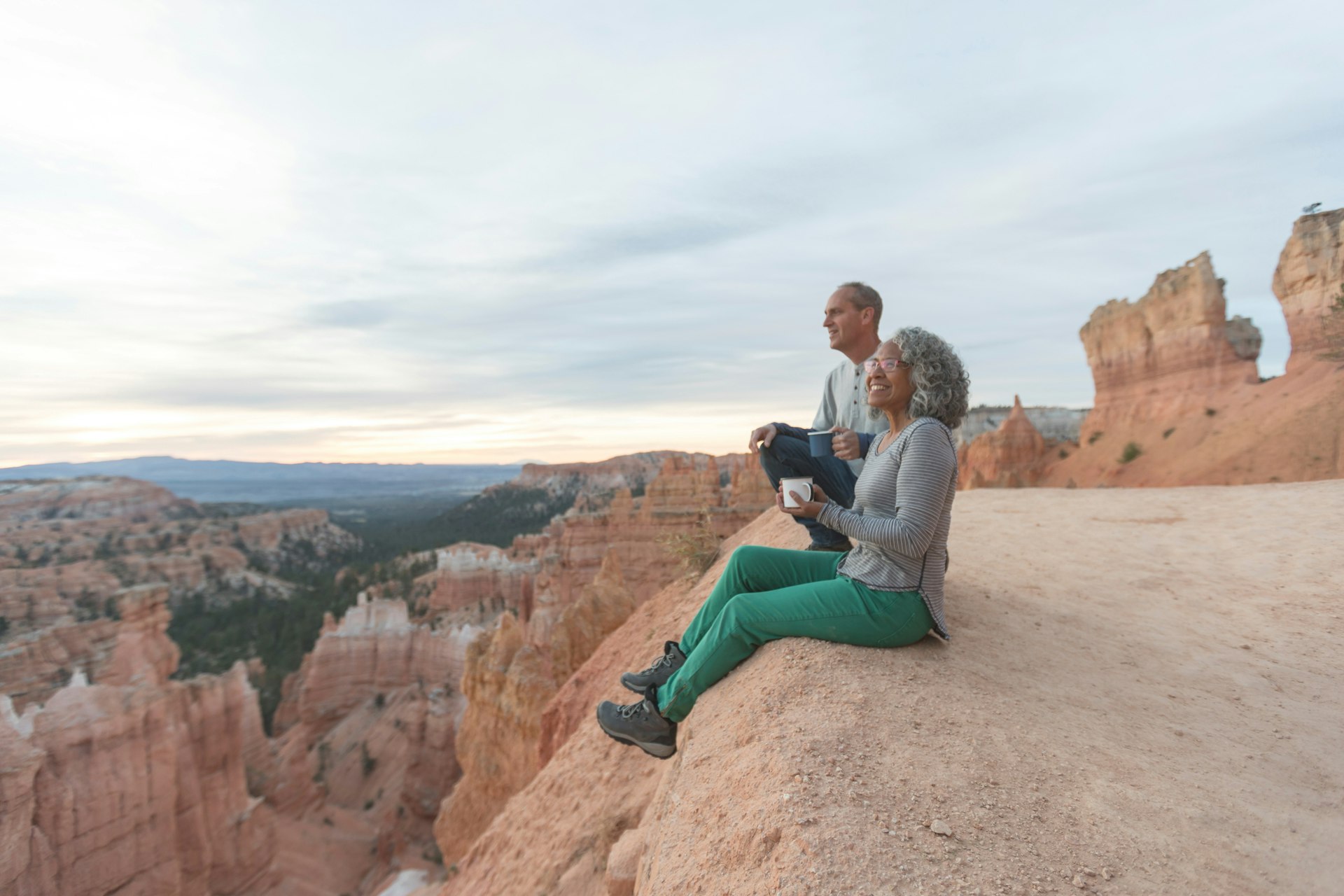 An older couple take a break from hiking to enjoy the view at Utah outlook overlooking a canyon. They are sitting on the cliff's edge and soaking in the scenery. The mountains and canyon are in front of them.