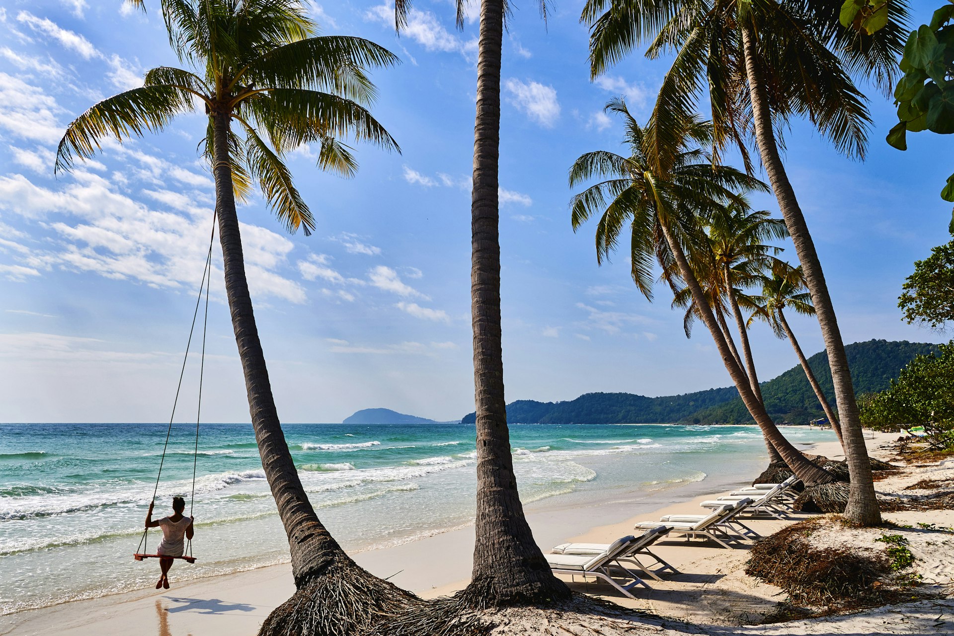 A woman sits on a swing connected to a palm tree on a beautiful beach