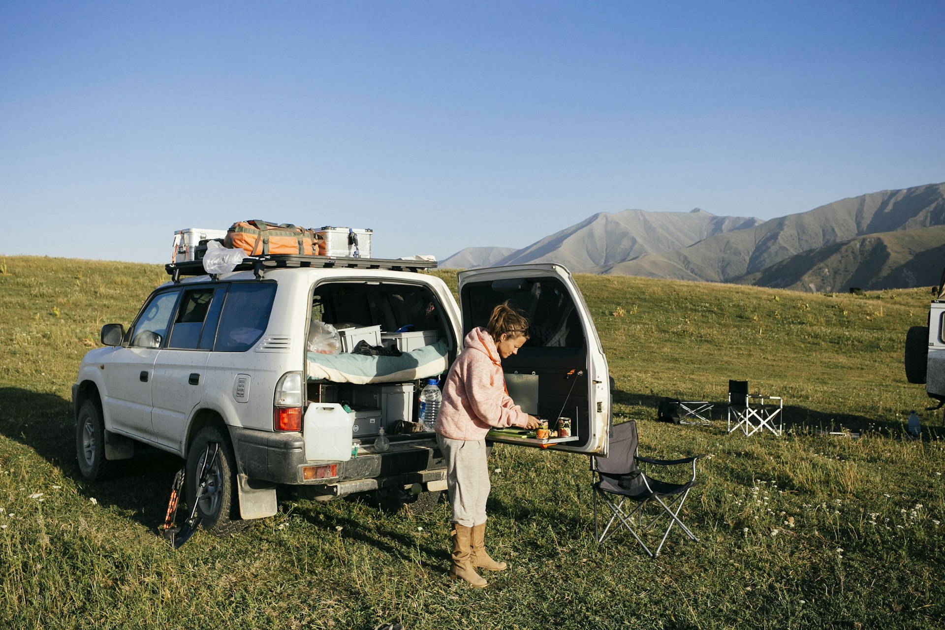A woman stands behind a fully packed 4WD prepping food in a meadow