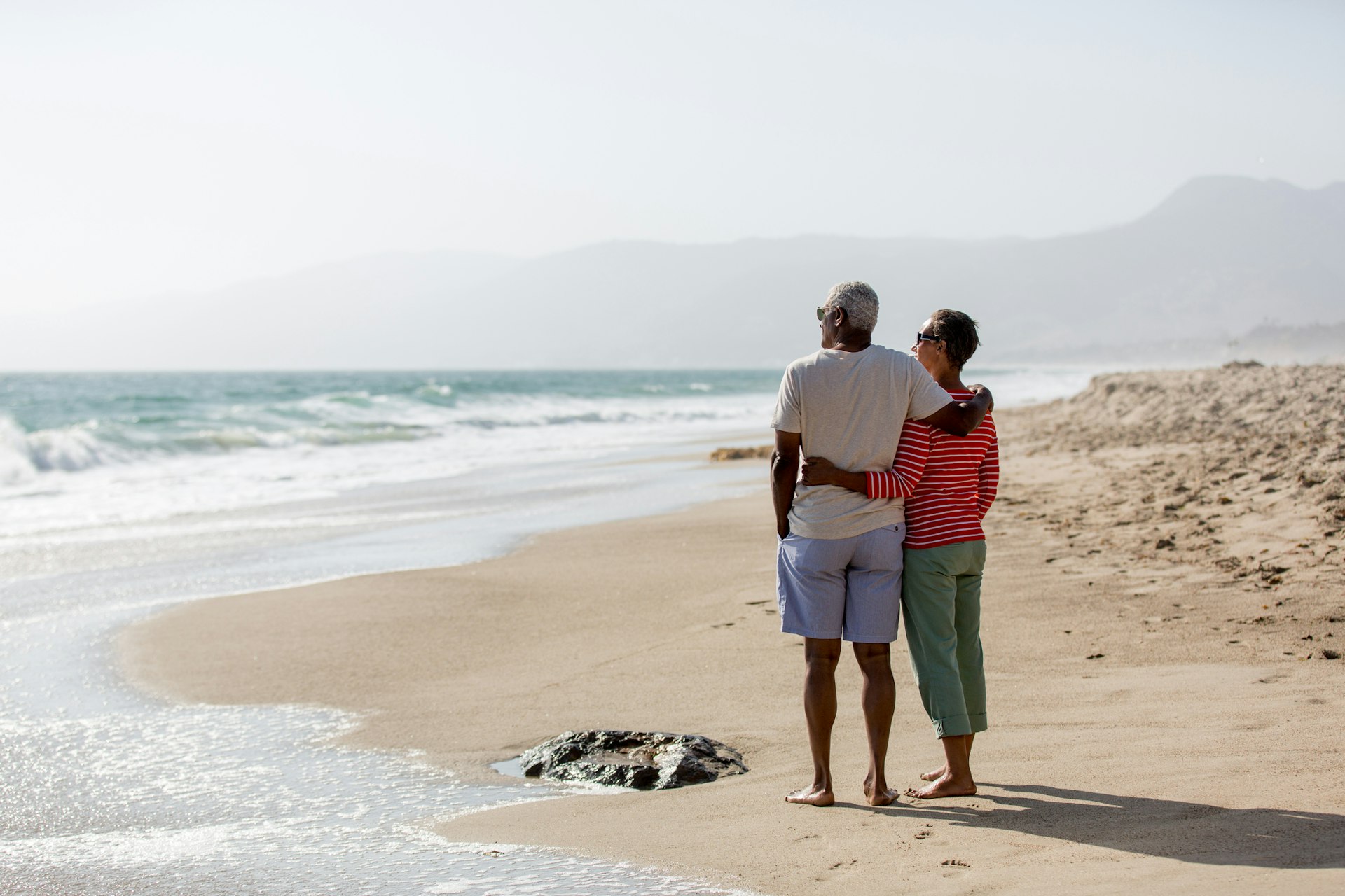A senior couple stands together on the sand looking out to sea