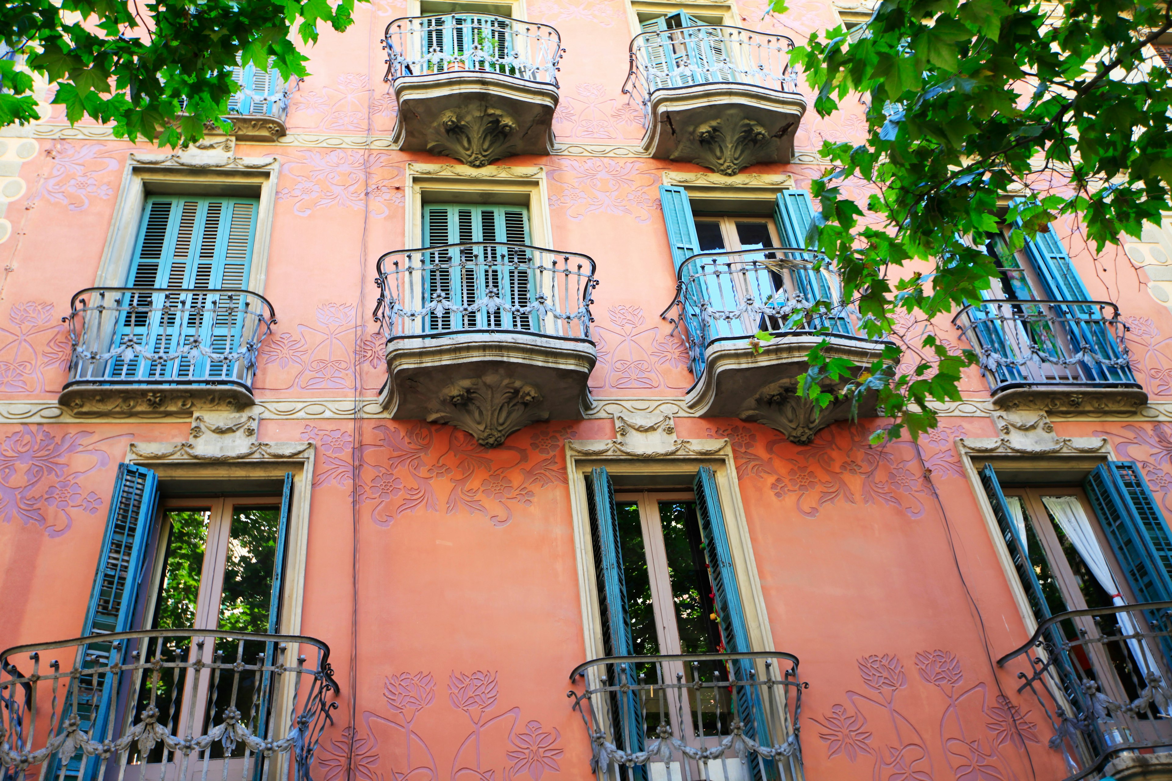 A red-walled building with blue shuttered windows and elaborate balconies; between each window is an intricate purple floral motif.