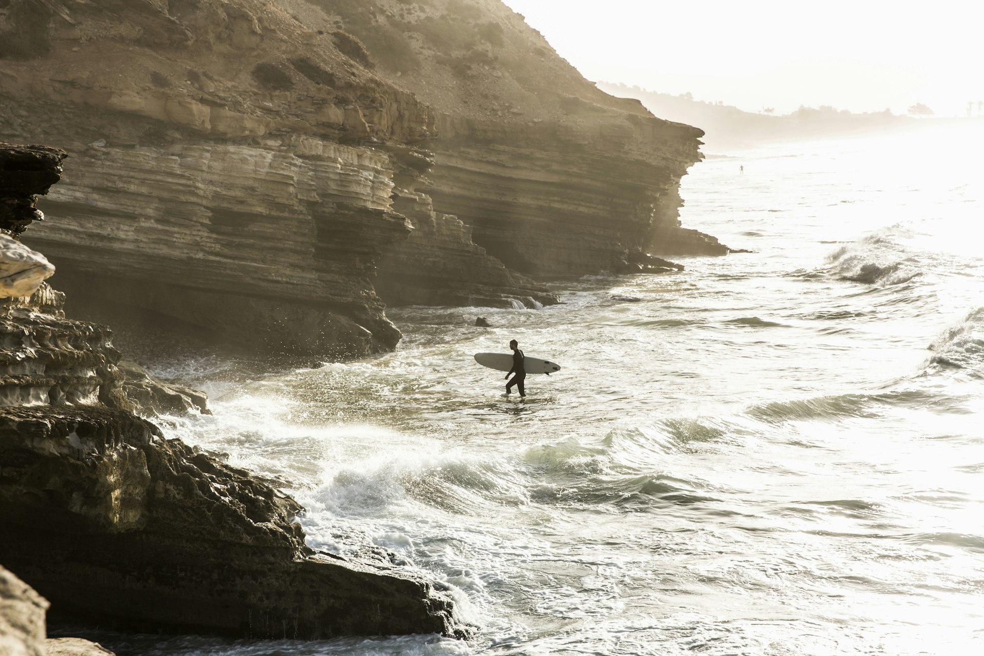 Surfer walks towards the rocky shore with his surfboard during sunrise, Taghazout Bay, Morocco