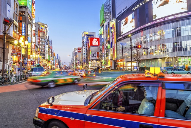 Tokyo taxi turning right in a busy road, traffic ahead has motion blur, the shops have neon billboards