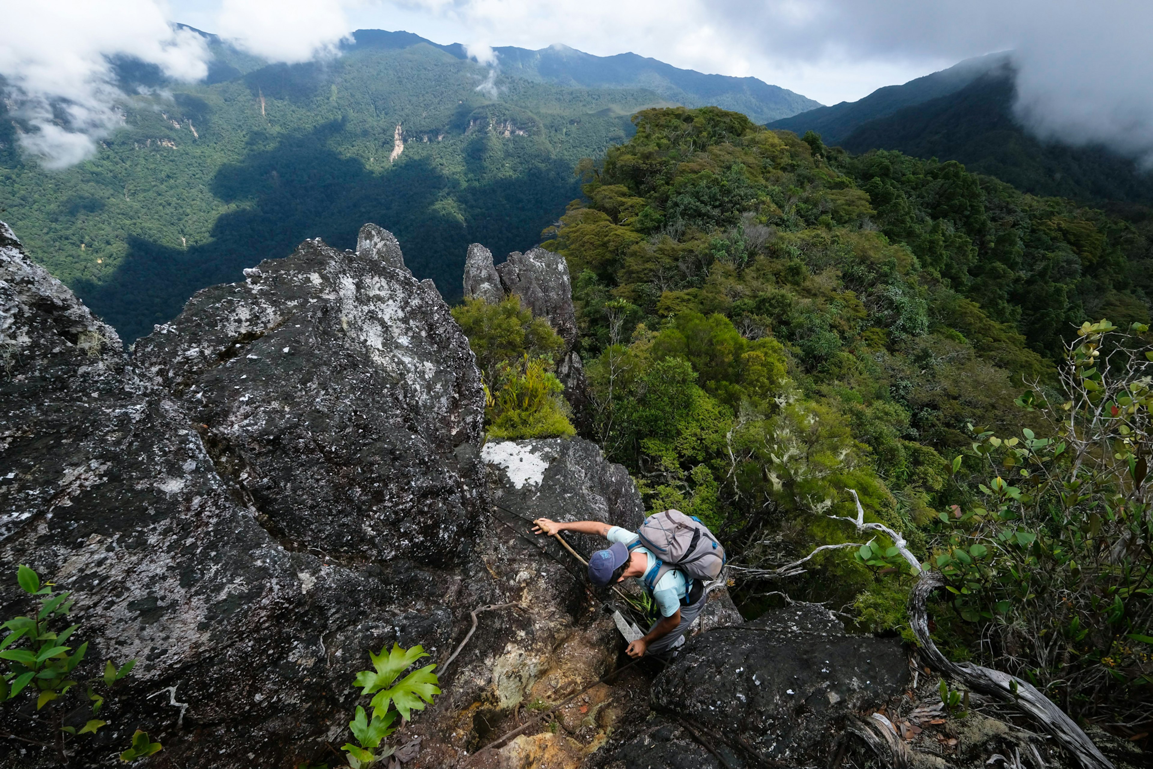A hiker seen from above scaling a rock face on a mountain covered with tropical vegetation, with wispy clouds in the distance
