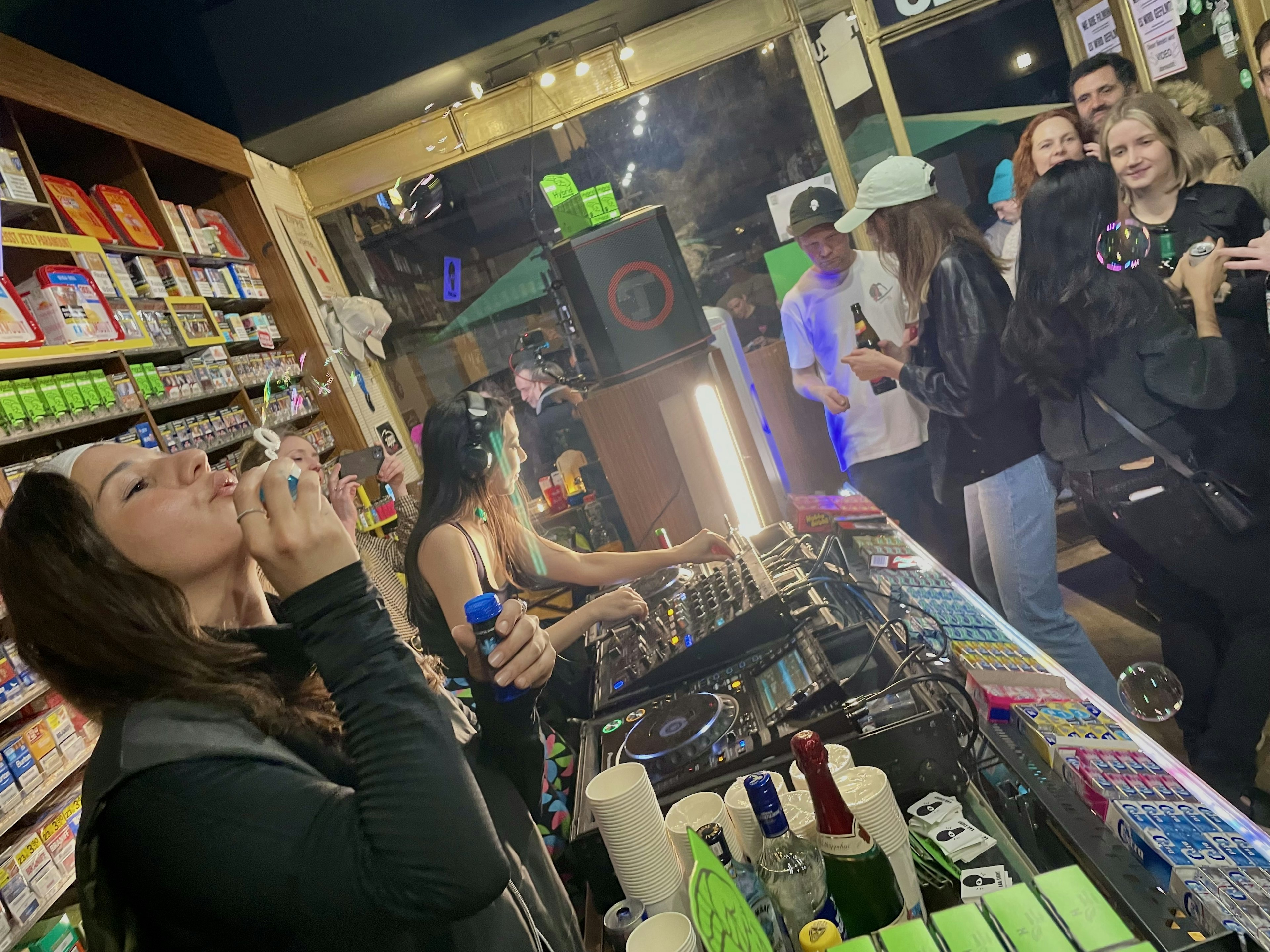 Two women djing in a Späti in Berlin while one blows bubbles